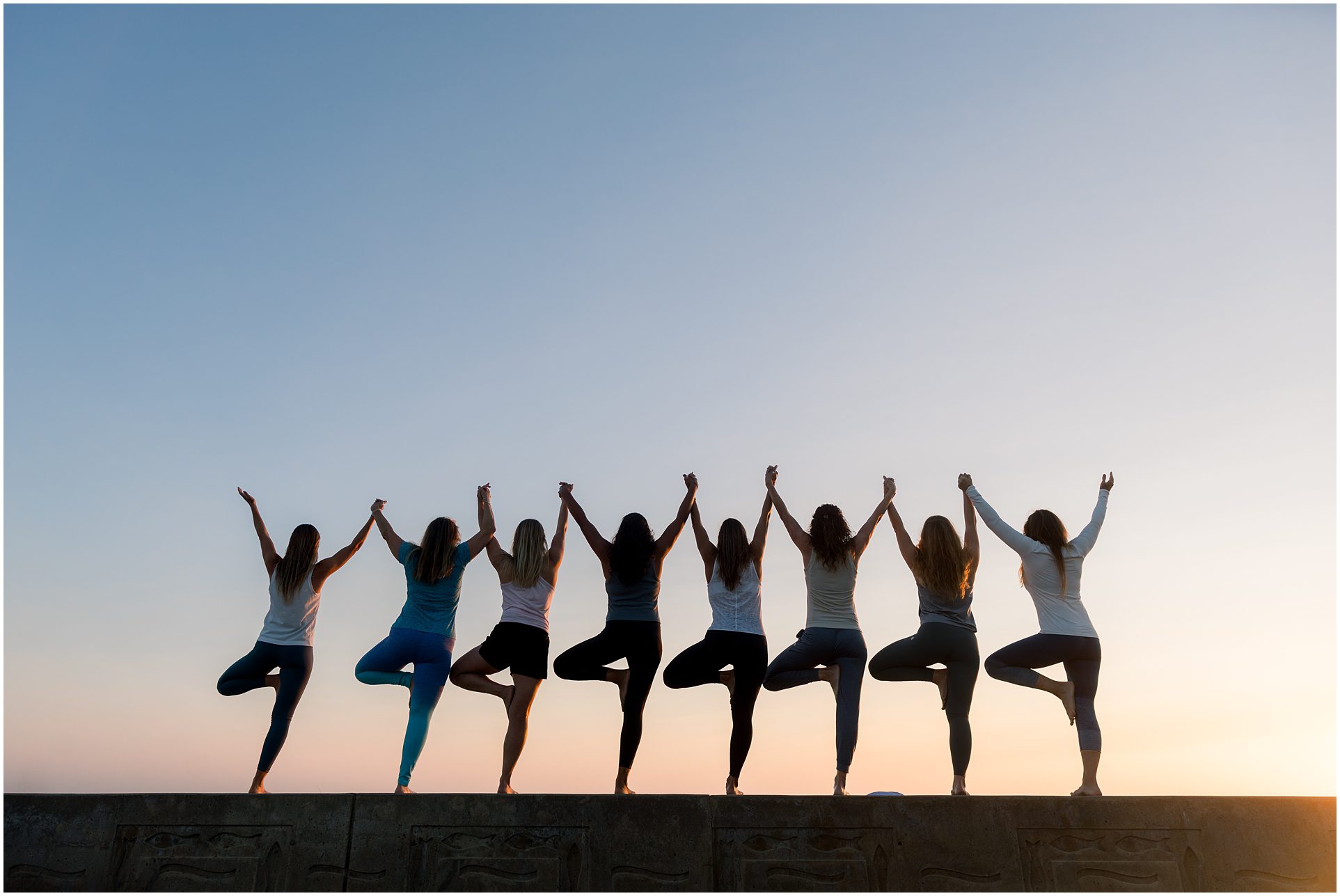 Eight people in workout clothes do tree pose on a ledge at sunset, holding hands with arms raised.