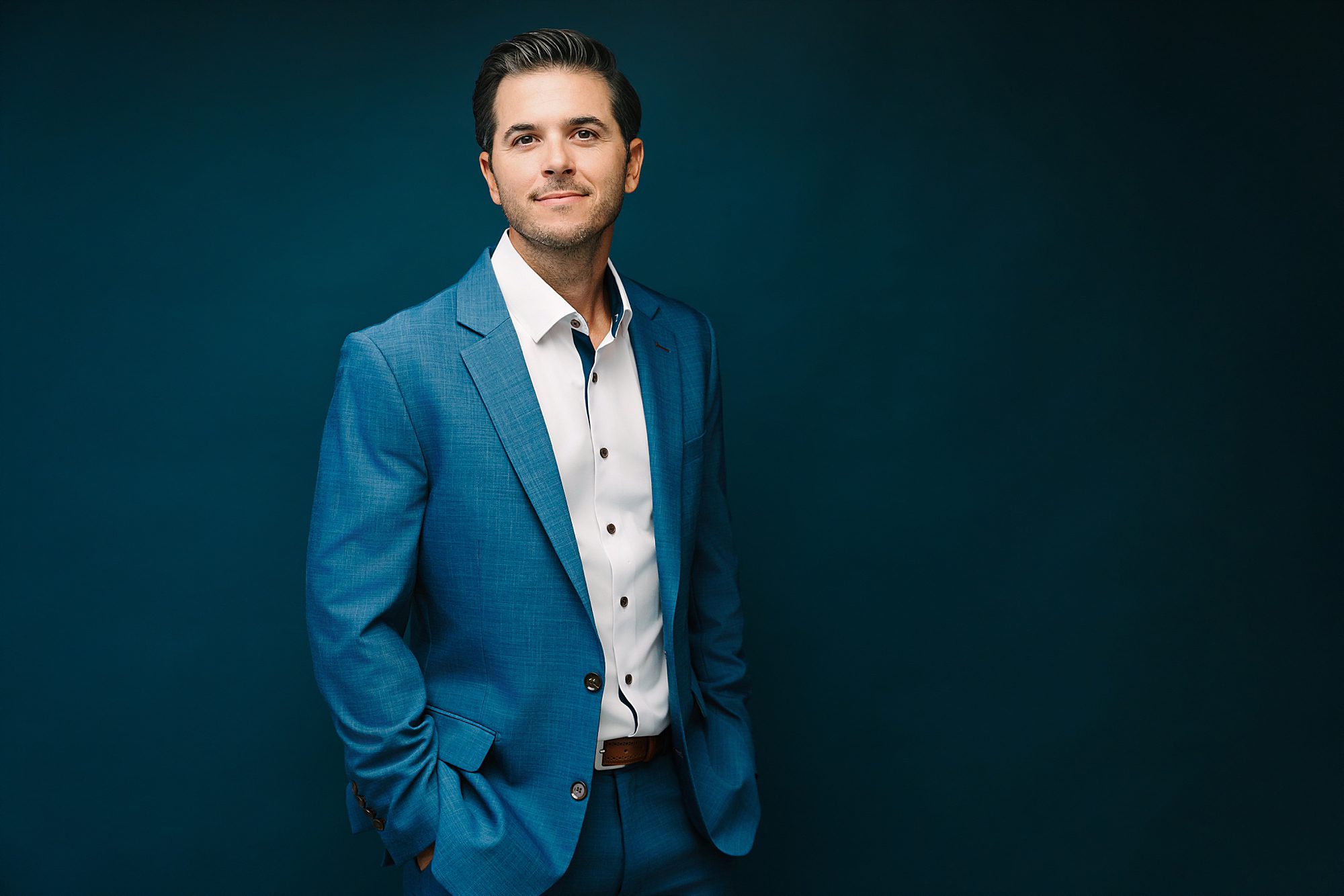 Headshot of Headshot of Man in a blue suit stands against a dark blue background, hands in pockets, smiling slightly.