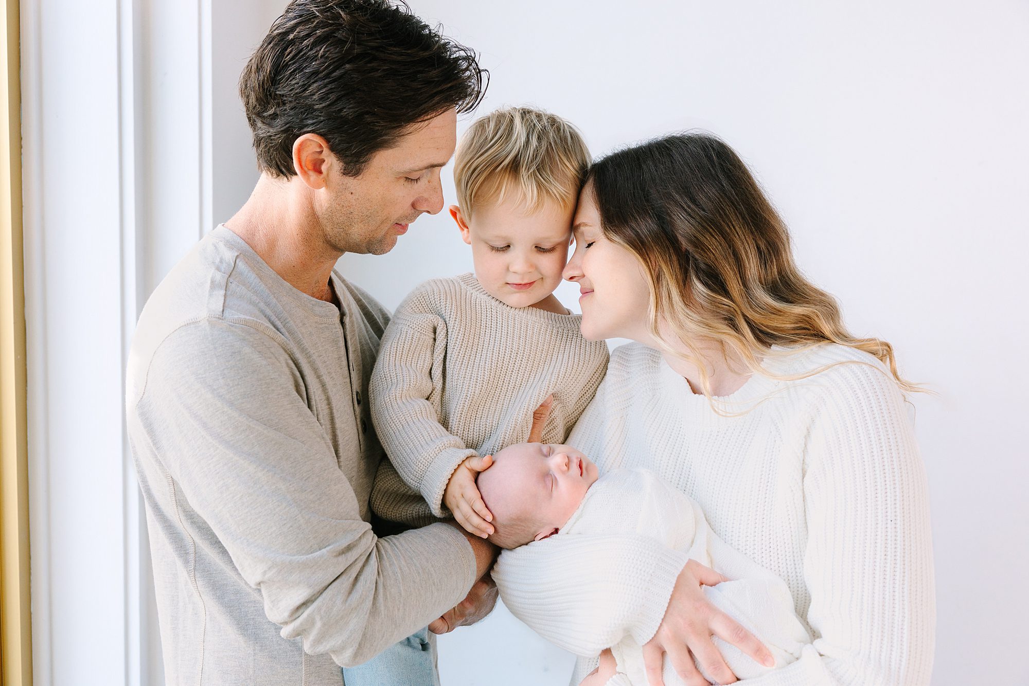 Parents and their toddler son look lovingly at a sleeping newborn baby held in their arms.