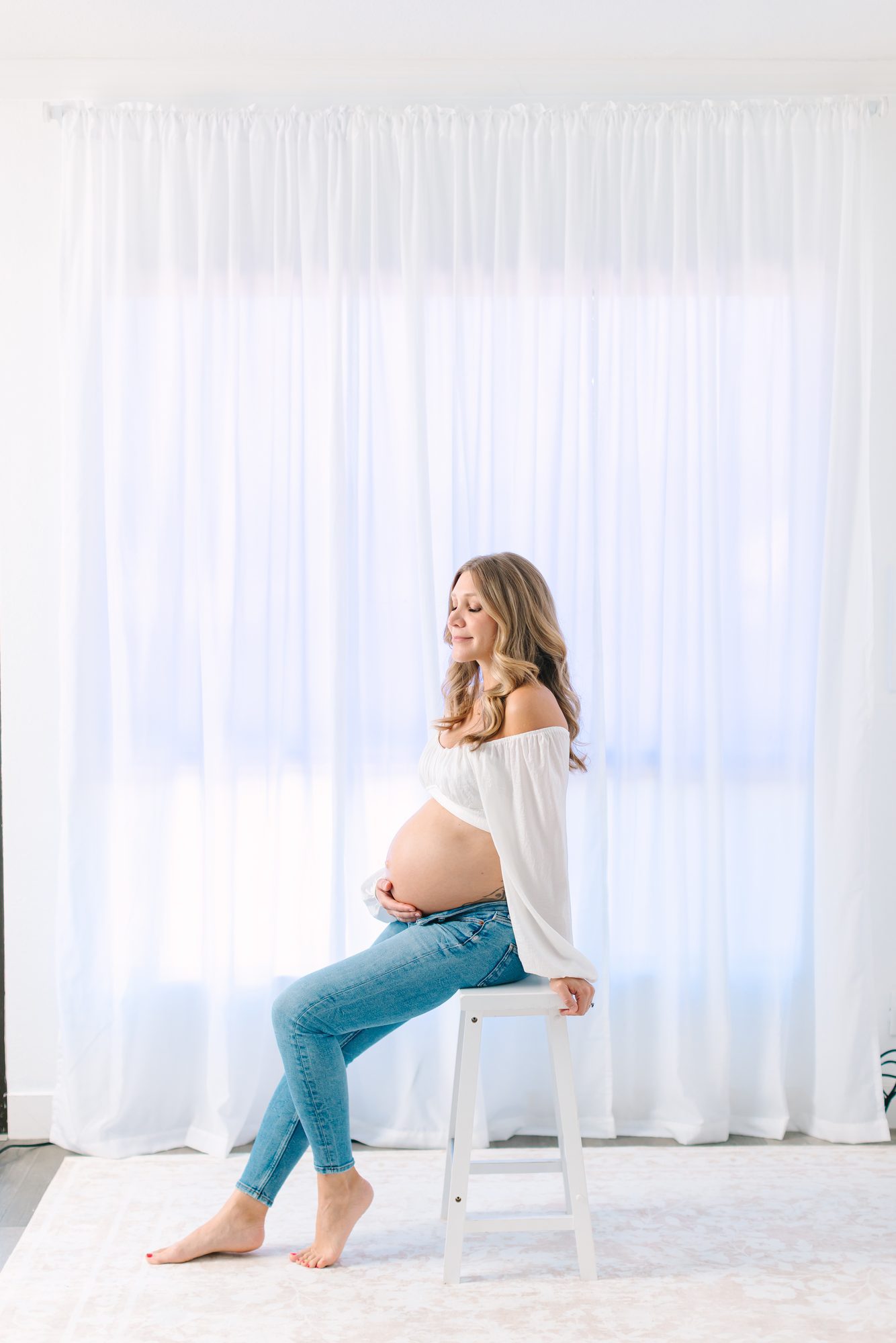 Expectant mother seated on a stool in front of sheer white curtains in the Carlsbad Village studio
