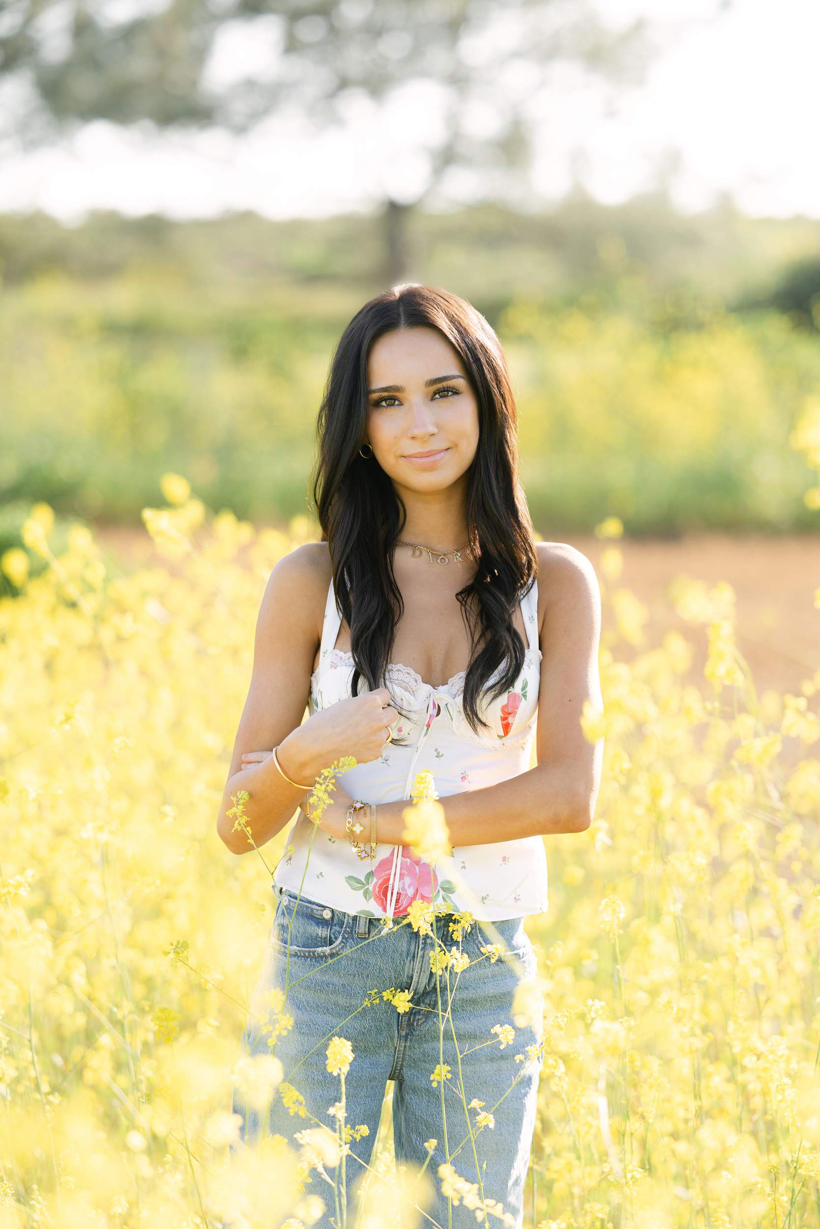 Young woman with long dark hair stands in a field of yellow flowers, wearing a floral top and jeans, smiling softly.