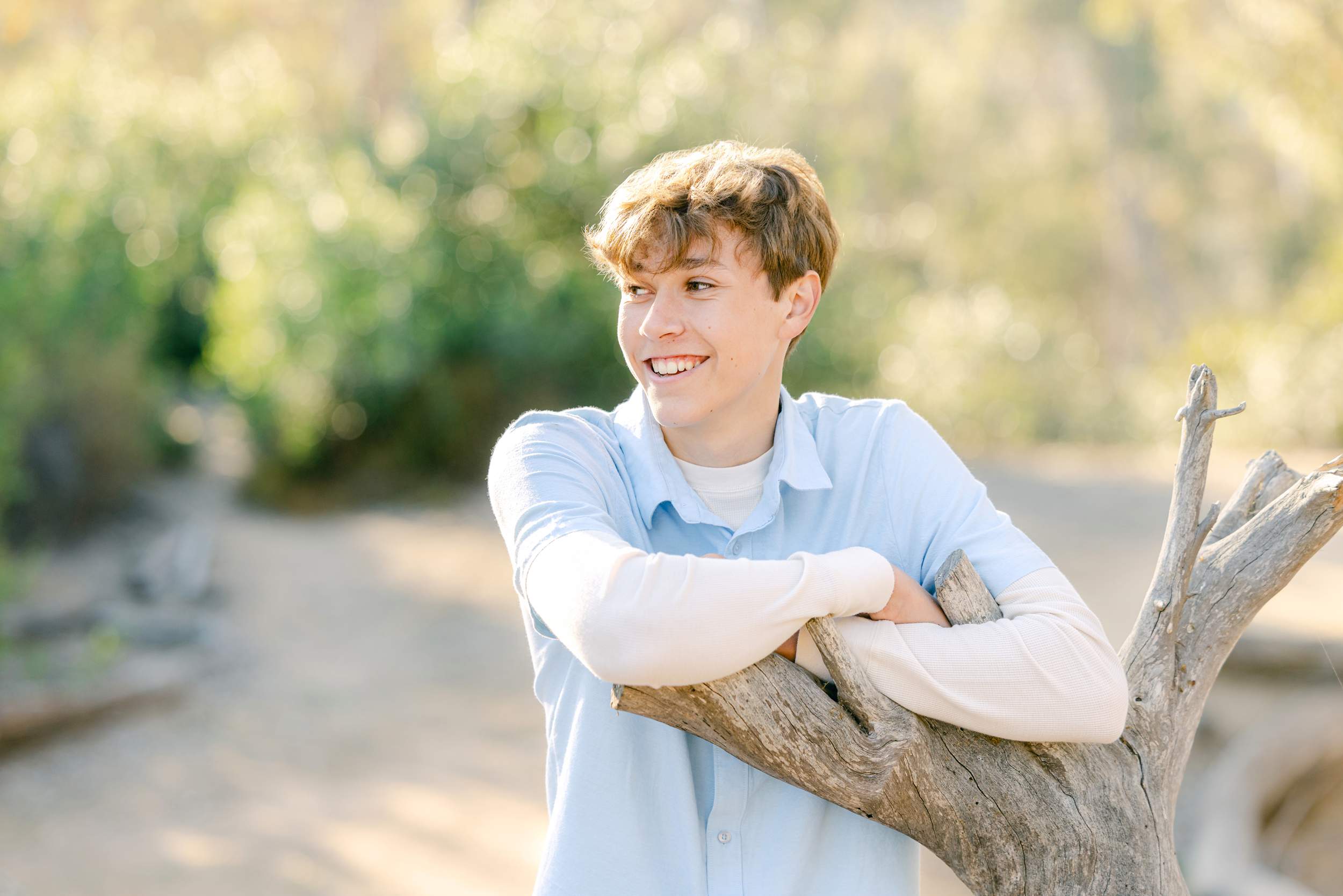 Young person in a light blue shirt leans on a tree branch outdoors, smiling and looking to the side.