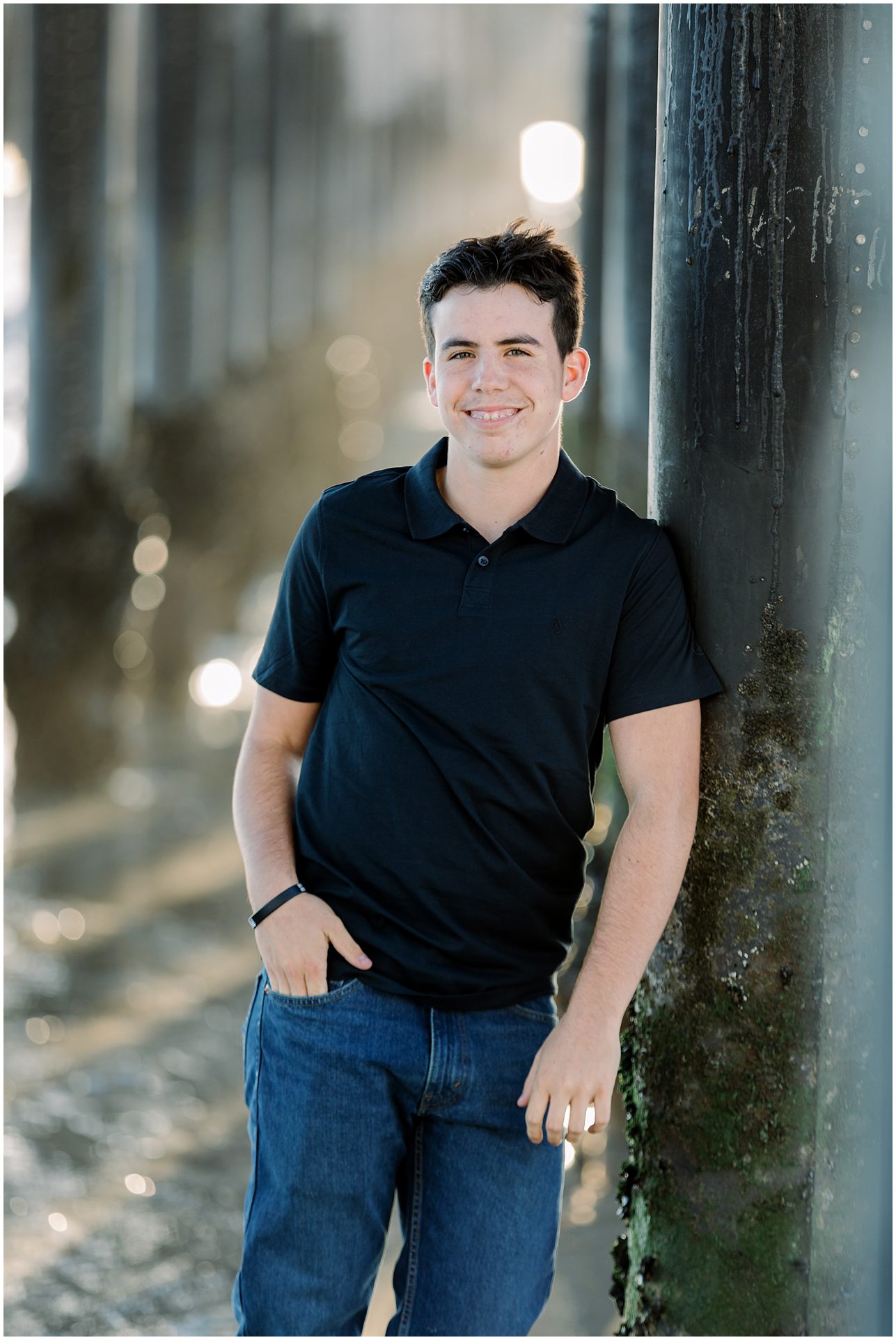 Young man in a black polo shirt leans against a pier column, smiling, with sunlight and water in the background.