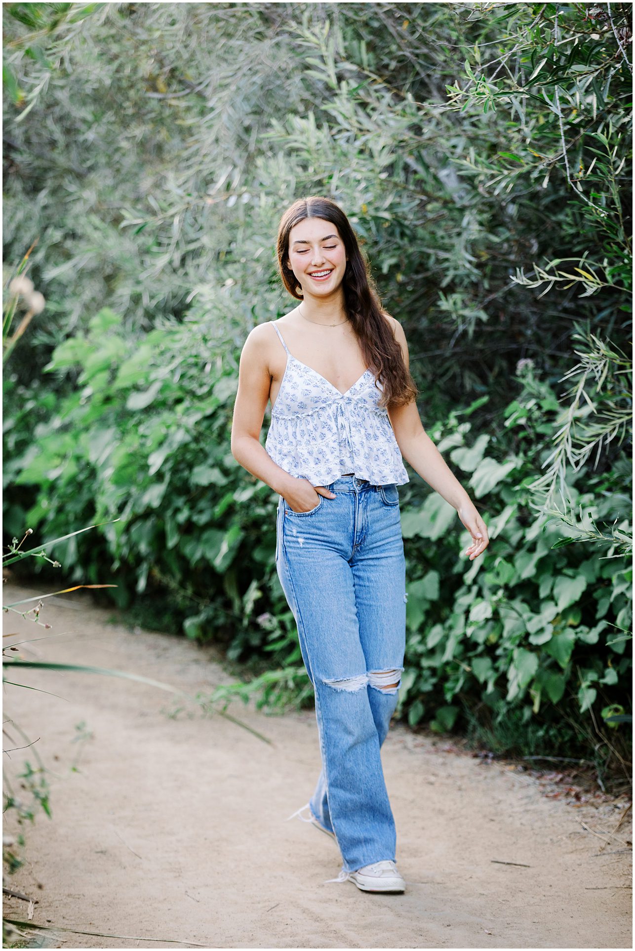 Young woman with long brown hair, wearing a tank top and ripped jeans, walks and smiles on a garden path.