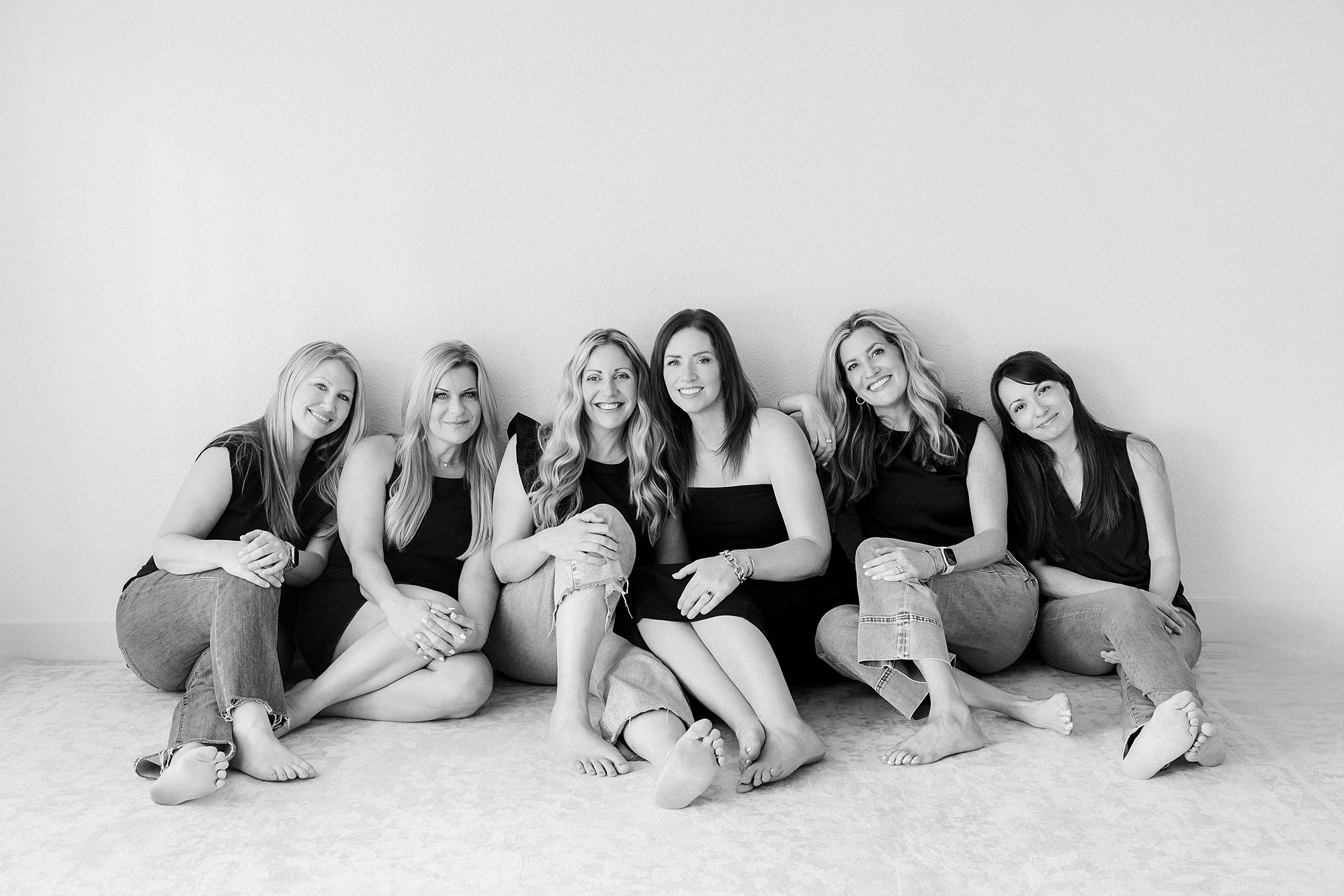 Six women sitting on the floor in a row against a plain wall.