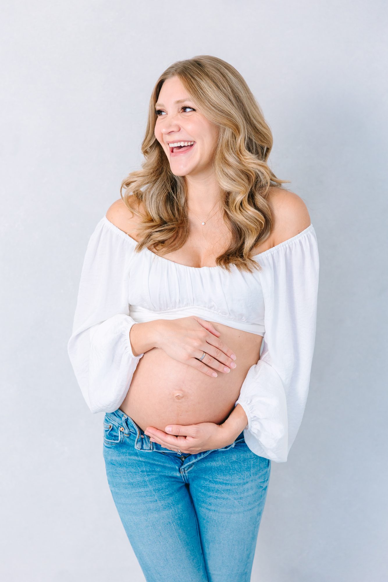 Laughing mother-to-be in a white top and jeans, studio maternity portrait