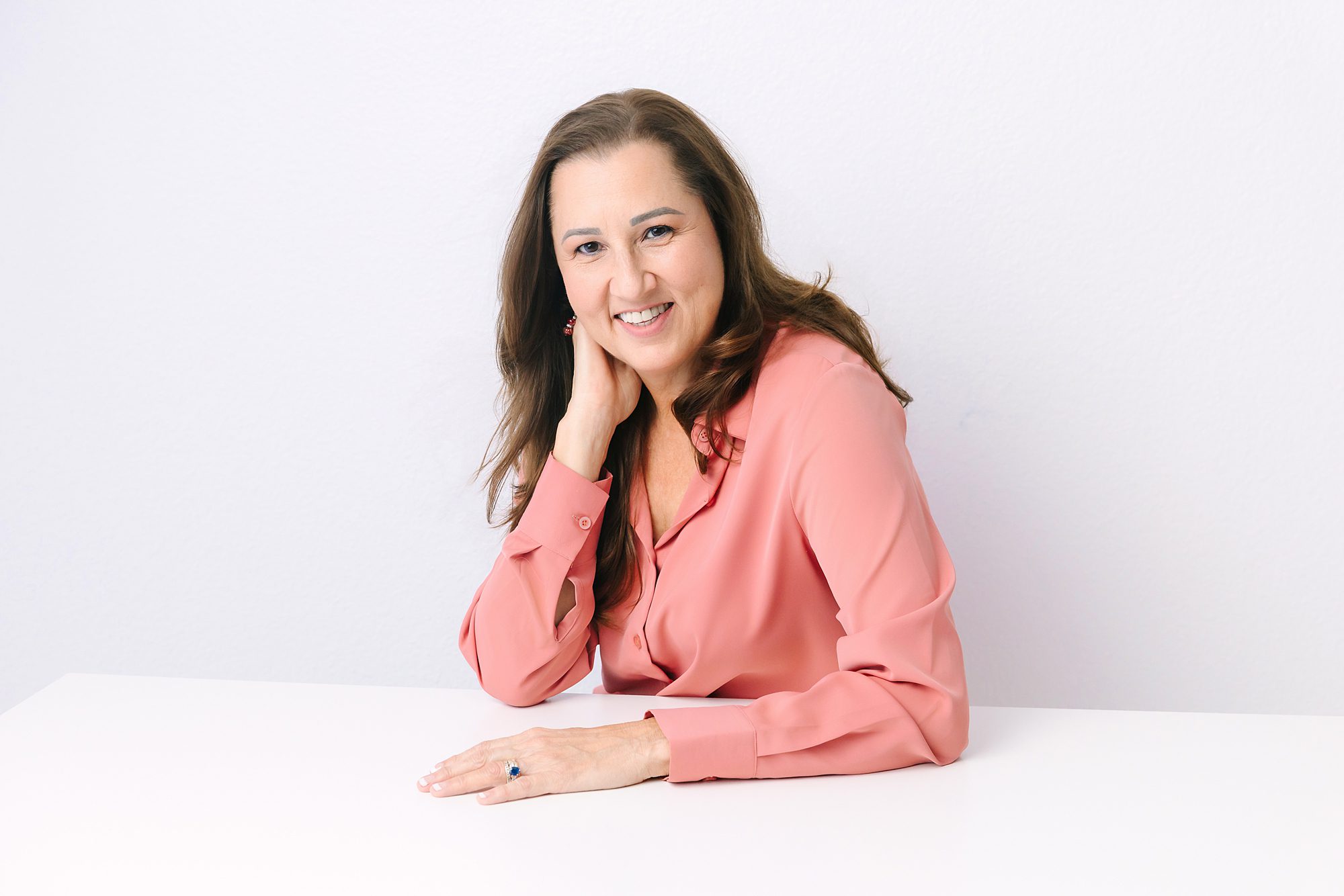 Headshot of Smiling woman in a pink blouse sits at a white table with her hand resting on her neck.
