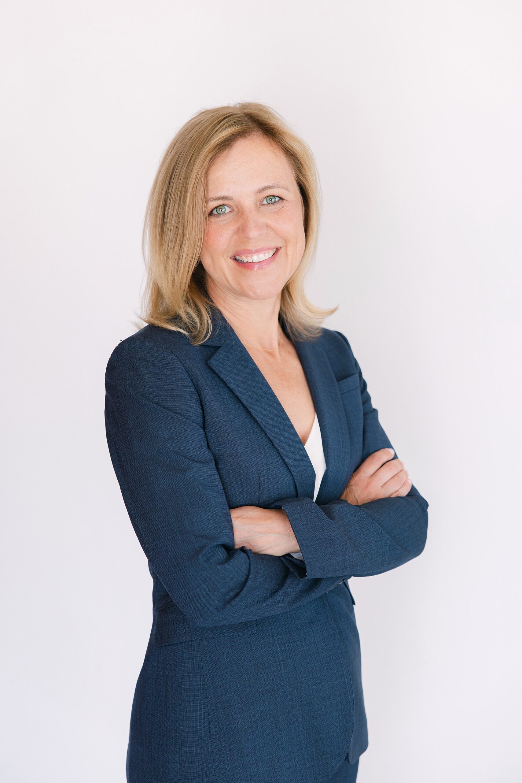 Headshot of Smiling woman with blonde hair in a blue suit stands with arms crossed against a plain white background.