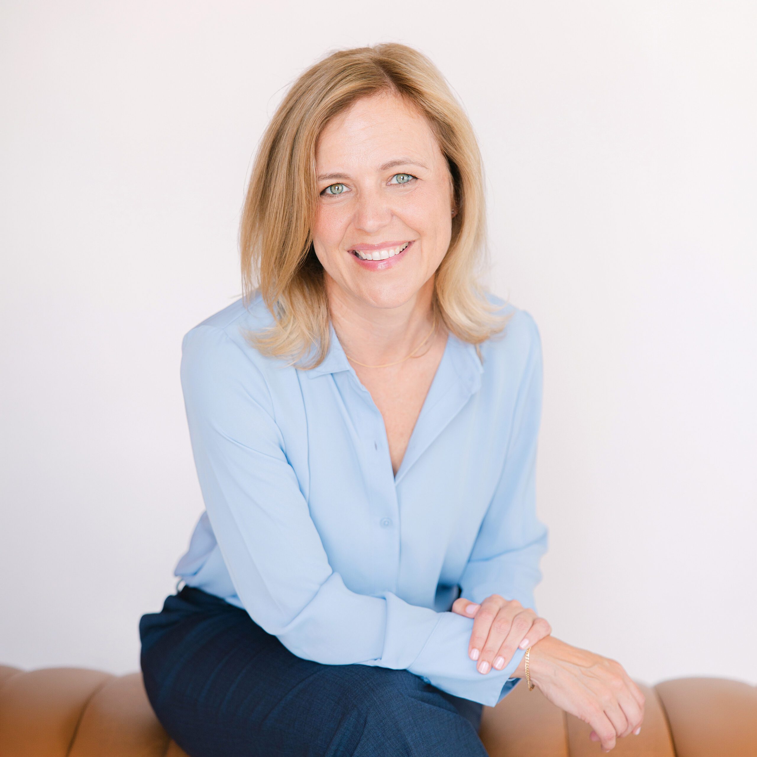 Carlsbad headshot of Smiling woman with blonde hair in a light blue blouse sits on a tan couch against a white background.