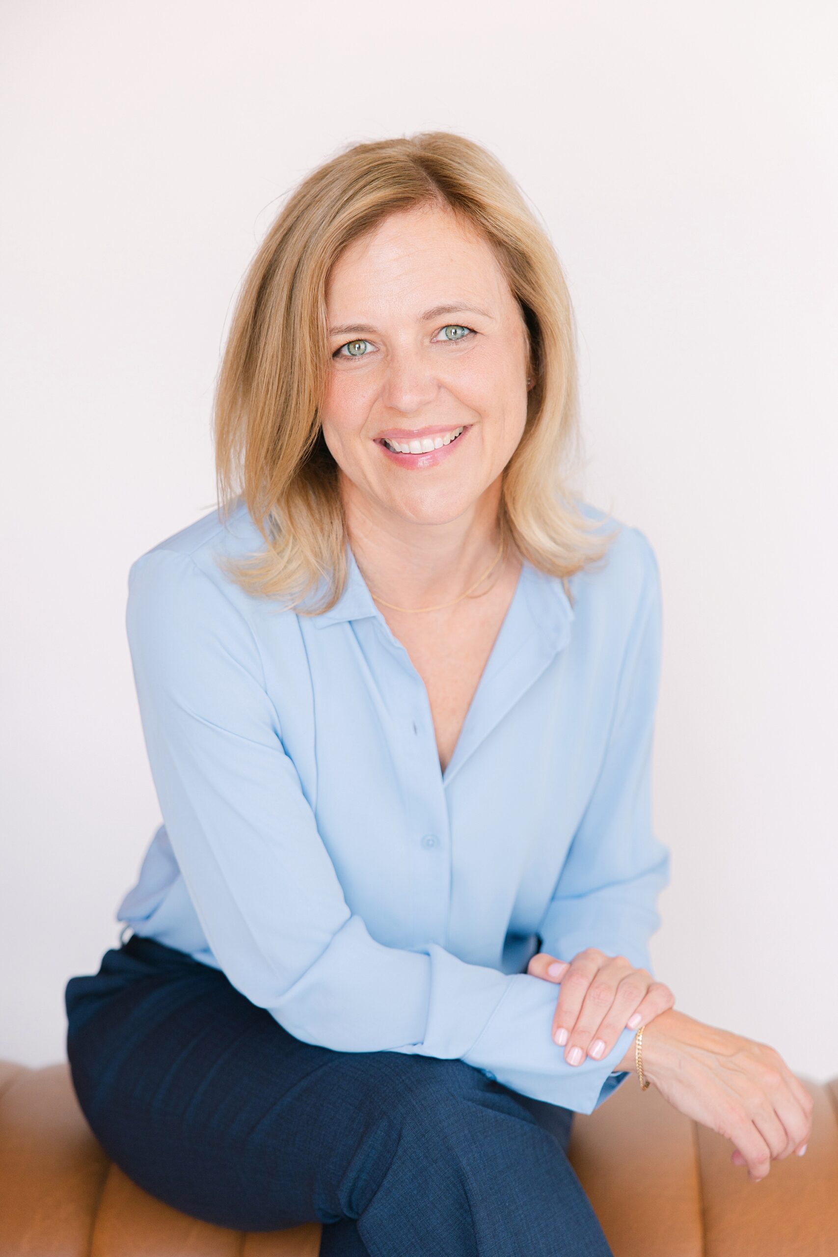 Headshot of Smiling woman with blonde hair, wearing a light blue blouse and dark pants, sitting on a tan chair.