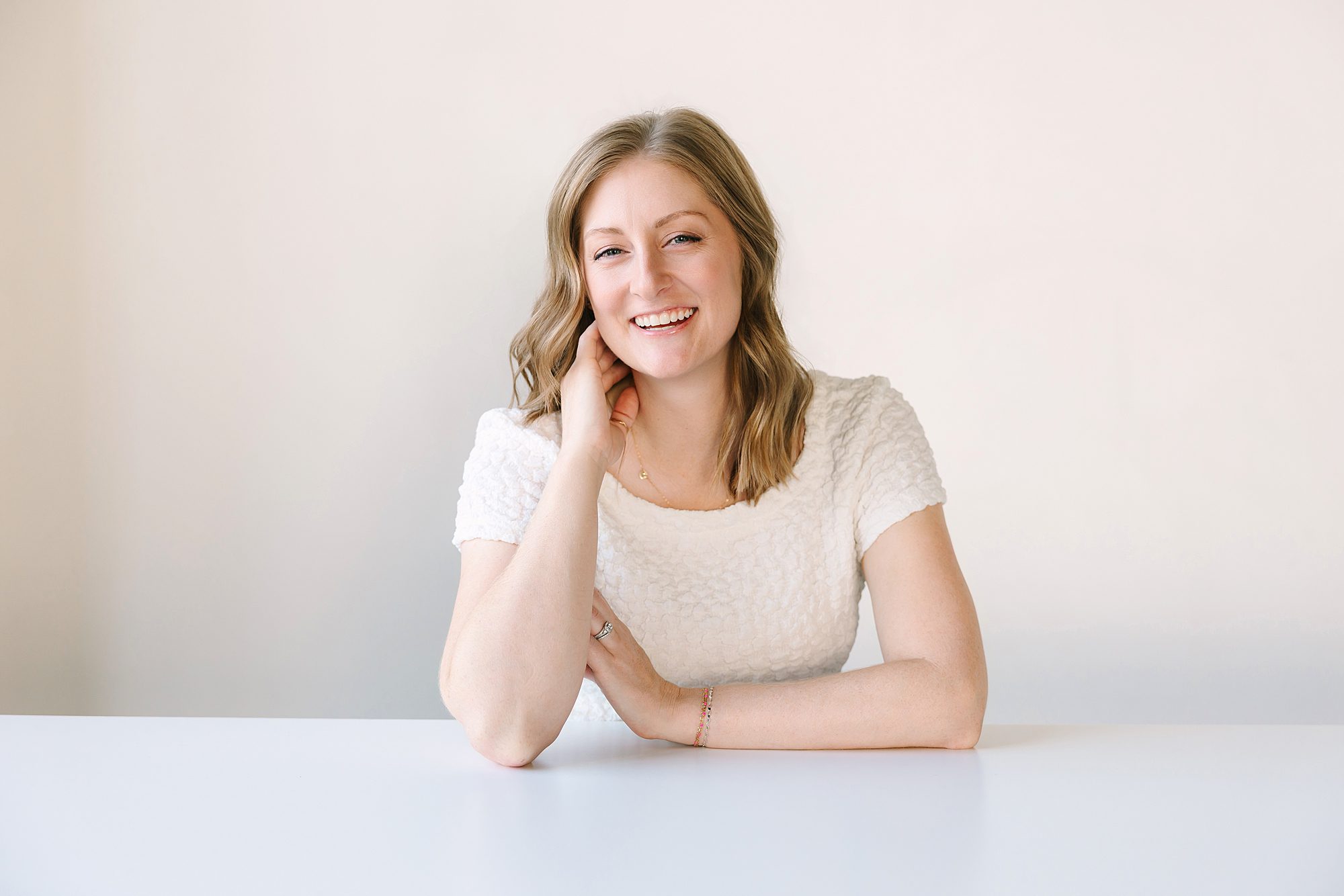 Headshot of Smiling woman with light brown hair in a white top sits at a white table against a plain background.