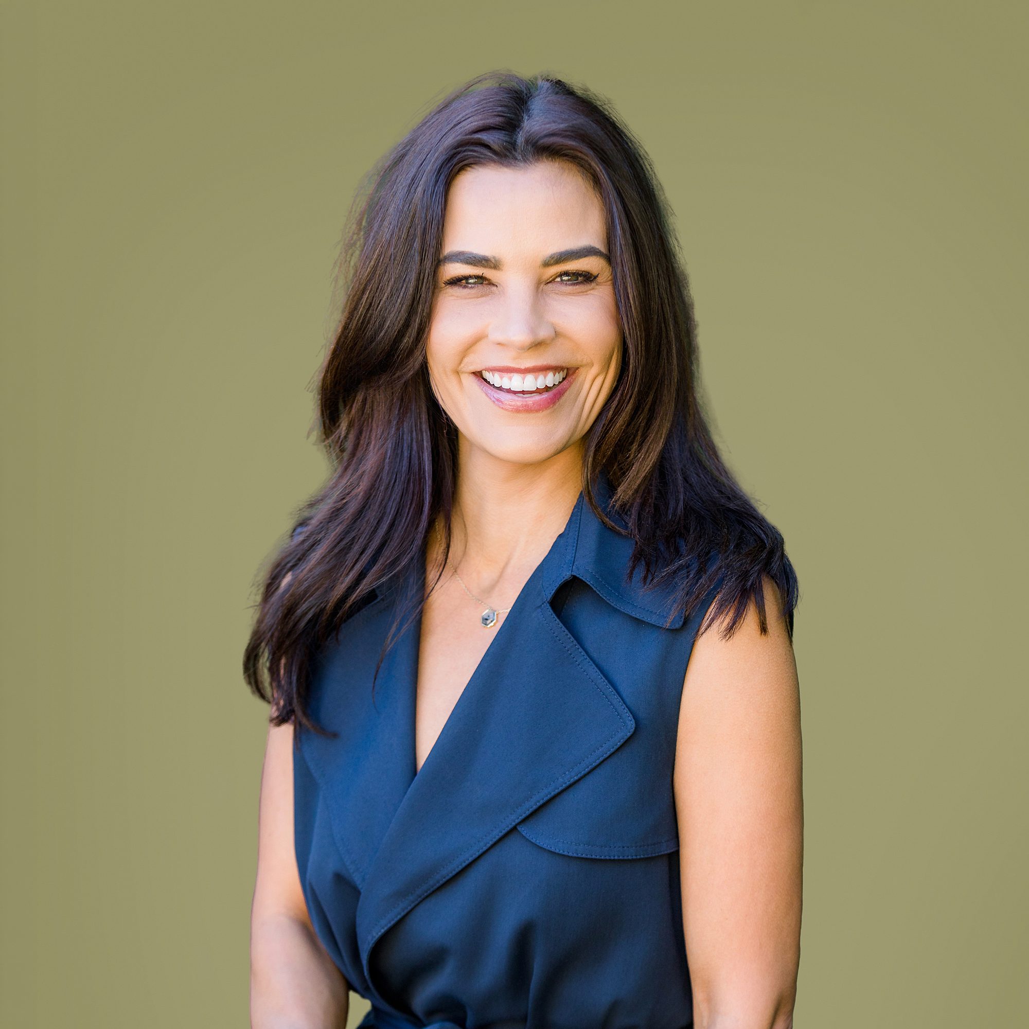Smiling woman with long brown hair in a sleeveless navy dress, standing against a plain olive background.
