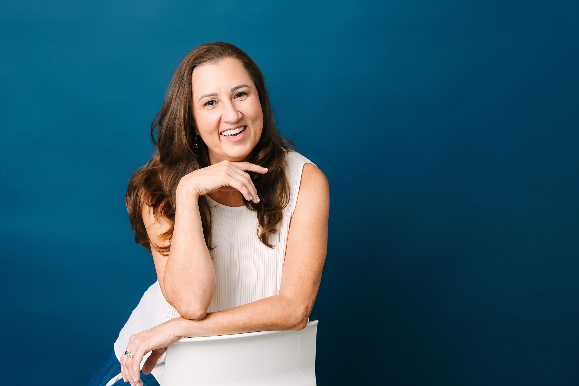 Headshot of Smiling woman with long brown hair sits on a white chair against a blue background, wearing a sleeveless white top.