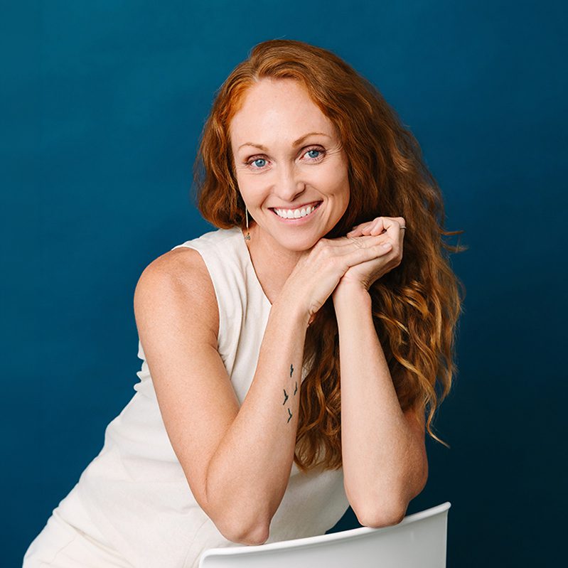 carlsbad headshot photographer Smiling with long red hair sits at a white chair against a blue background, hands resting on chair back.