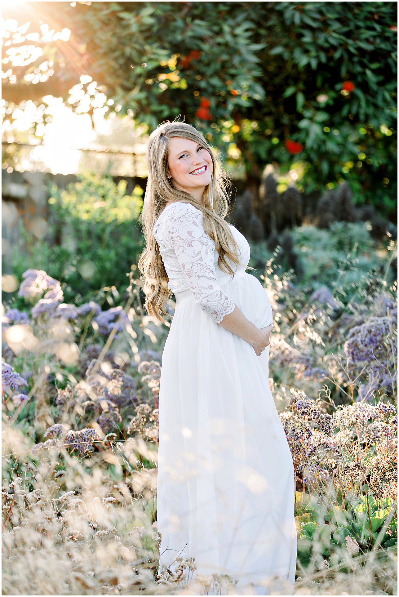 Pregnant woman in a white dress standing in a sunny garden, smiling and holding her belly.