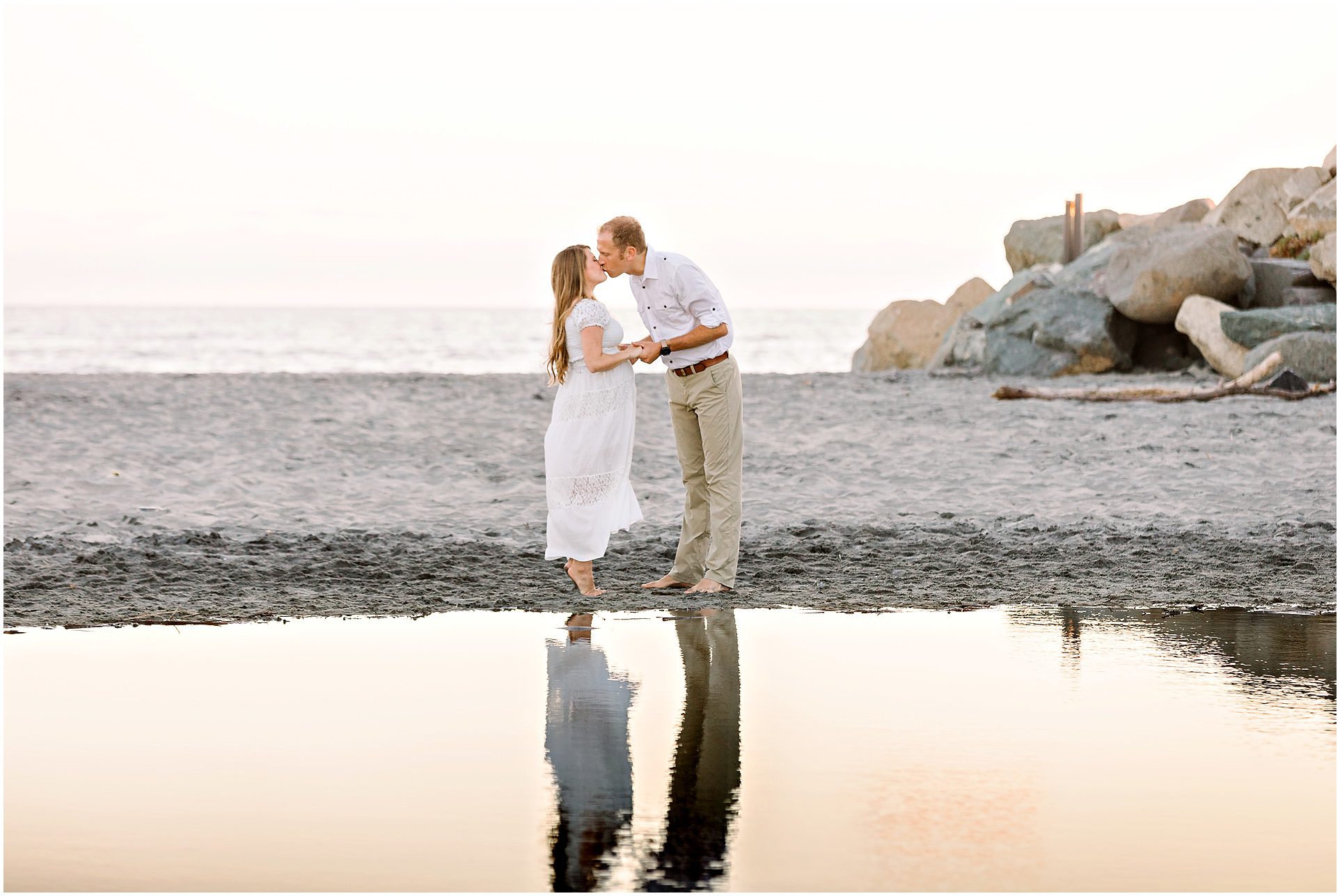 A couple in white clothes kisses on a sandy beach near rocks, reflected in calm water at sunset.