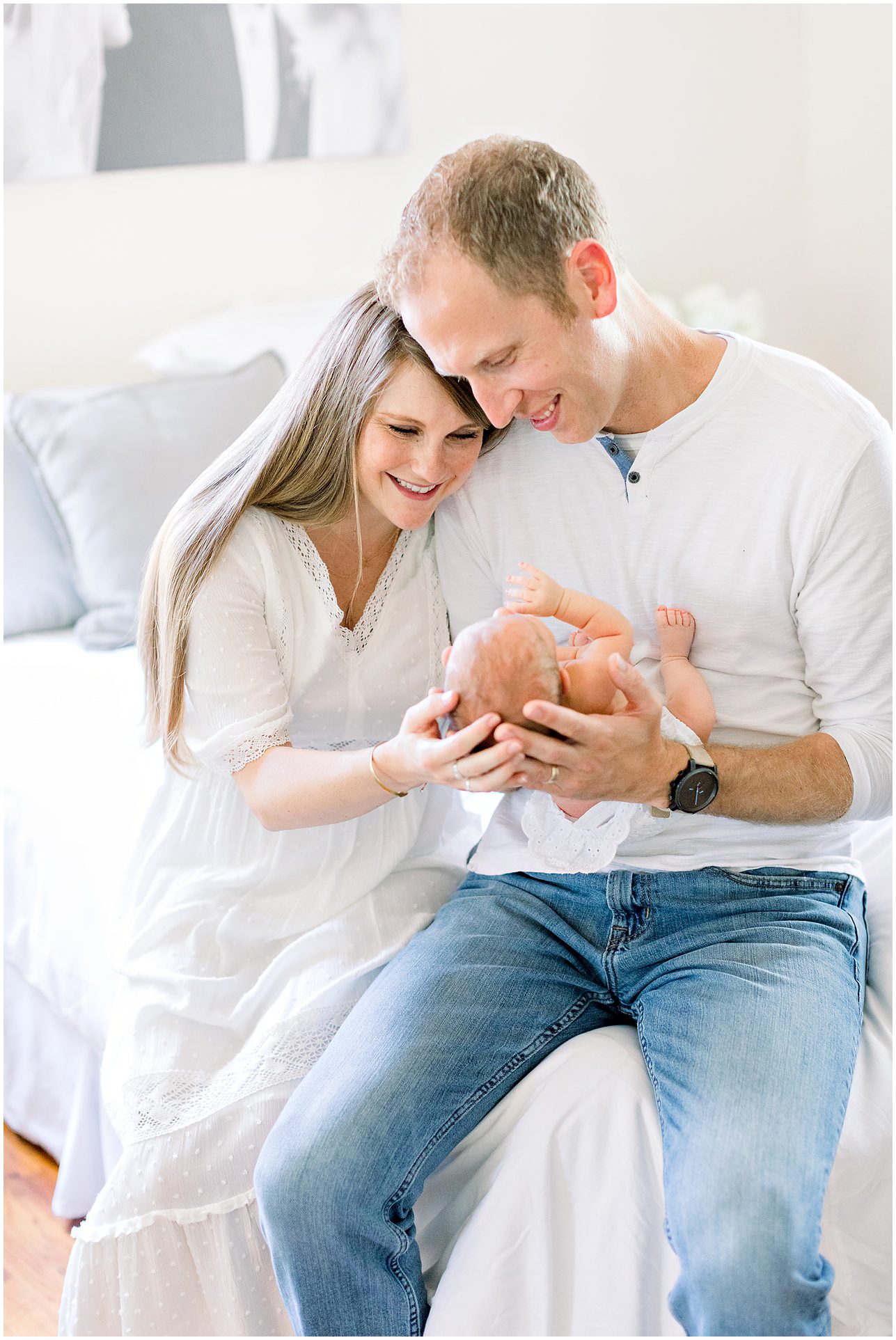 Smiling couple in white clothes sitting on a bed, holding and looking lovingly at their newborn baby.
