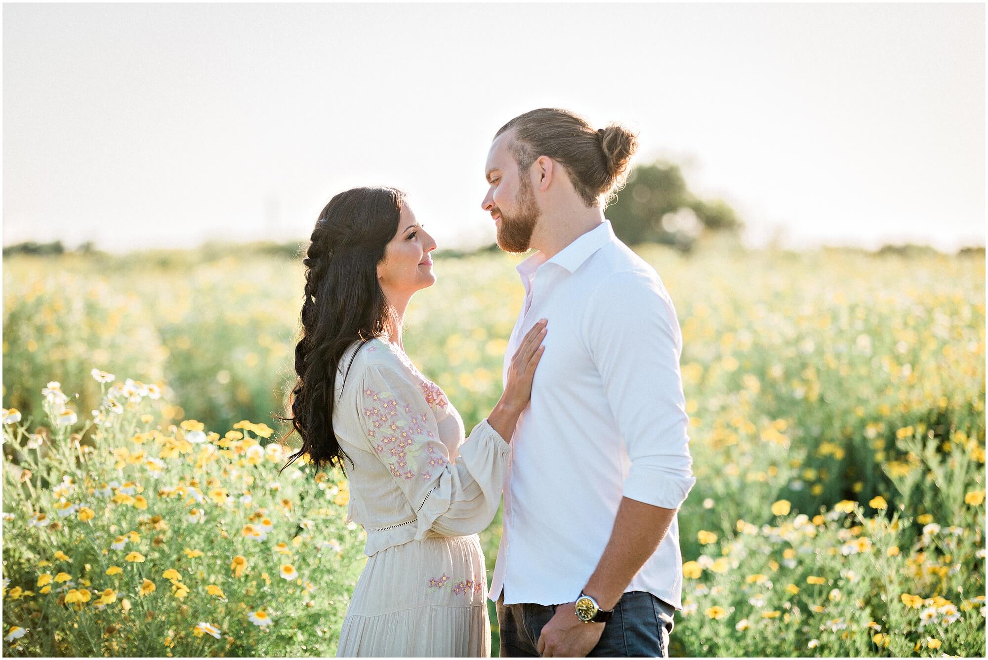 A couple stands close together in a sunlit field of yellow flowers, gazing lovingly into each others eyes.