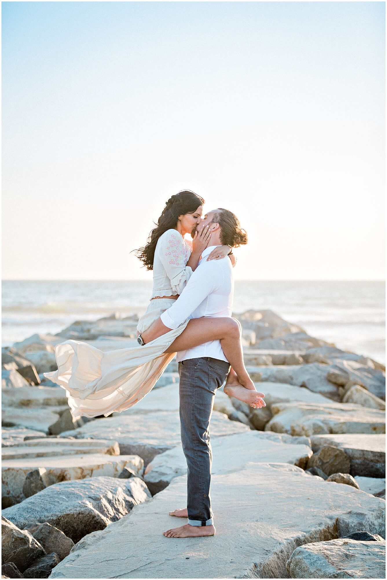 A couple kisses on a rocky pier by the ocean; the woman is lifted in the mans arms, her dress flowing in the breeze.
