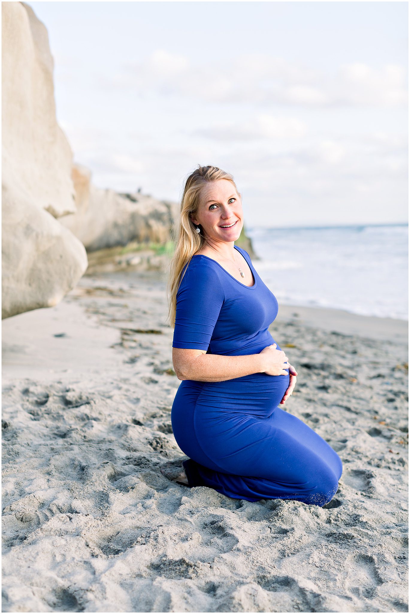 Pregnant woman in a blue dress kneeling on a sandy beach, smiling at the camera with the ocean in the background.