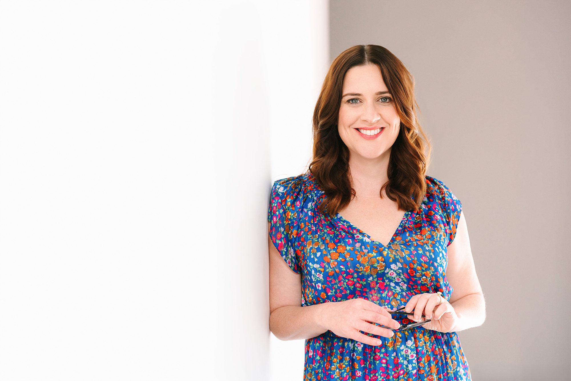 Headshot of Woman in a blue floral dress smiling, standing against a white wall, holding glasses in her hands.