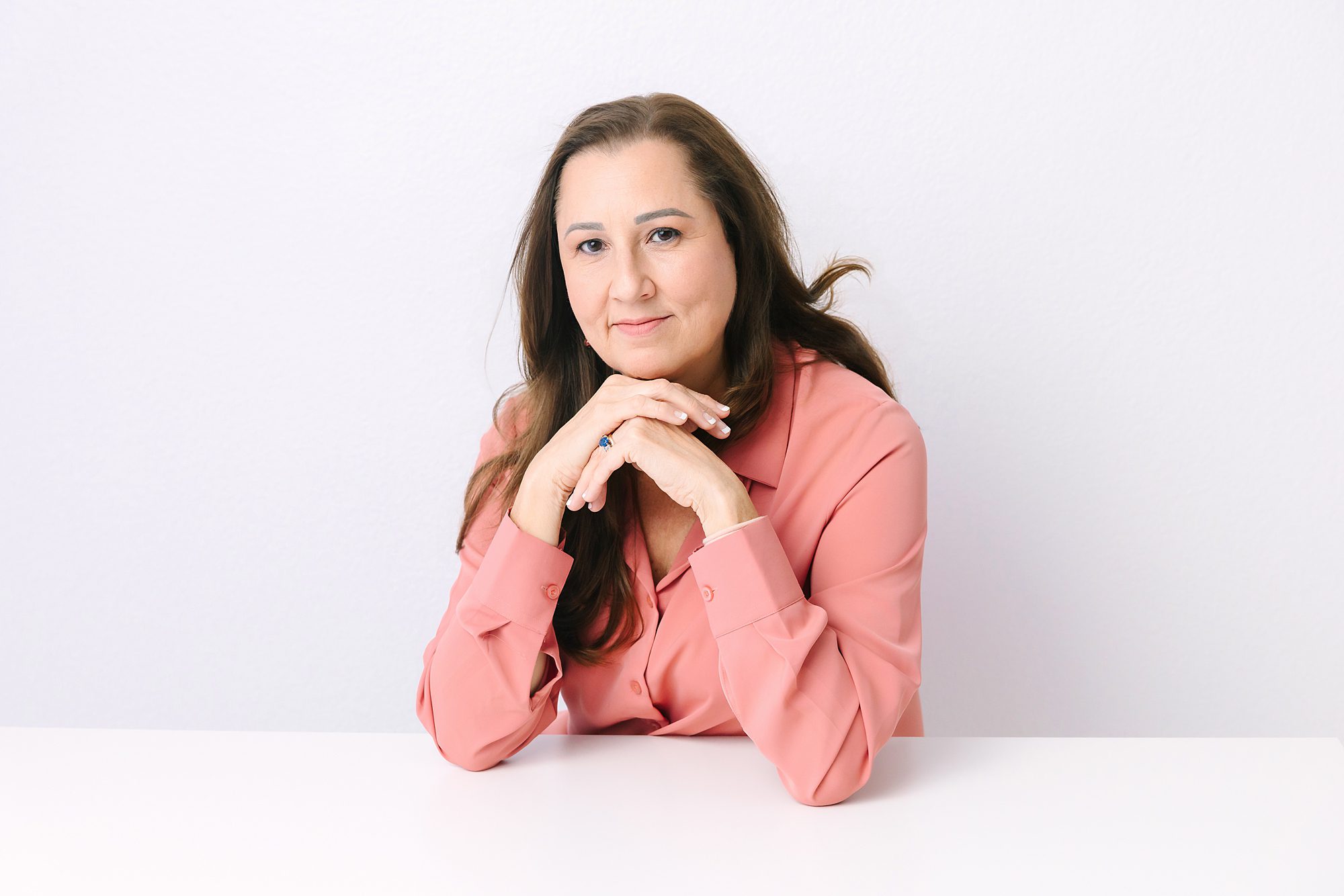 Headshot of Woman in a pink shirt sits at a white table with her hands folded, smiling softly at the camera.