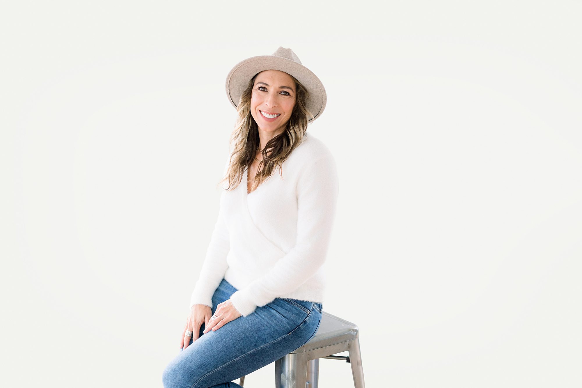 Headshot of Woman in a white sweater and hat sits on a stool, smiling, against a plain white background.