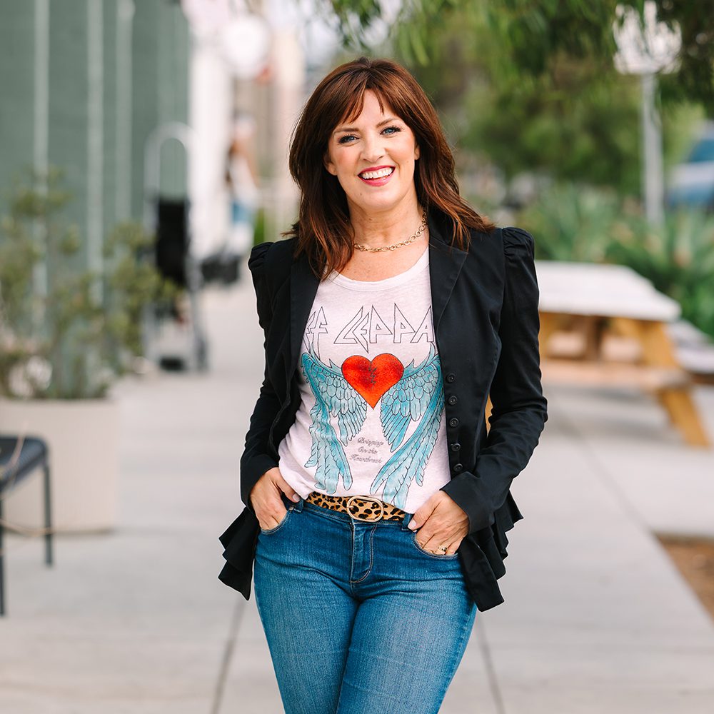 Carlsbad personal branding photo of Woman smiling outdoors, wearing a black jacket, graphic tee with wings and heart, and blue jeans.
