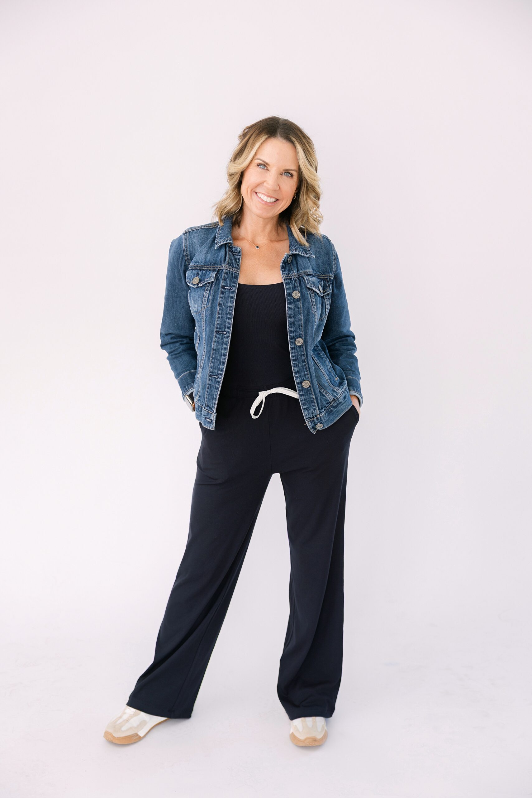 Headshot of Woman smiling, wearing a denim jacket, black jumpsuit, and sneakers, standing with hands in her pockets.