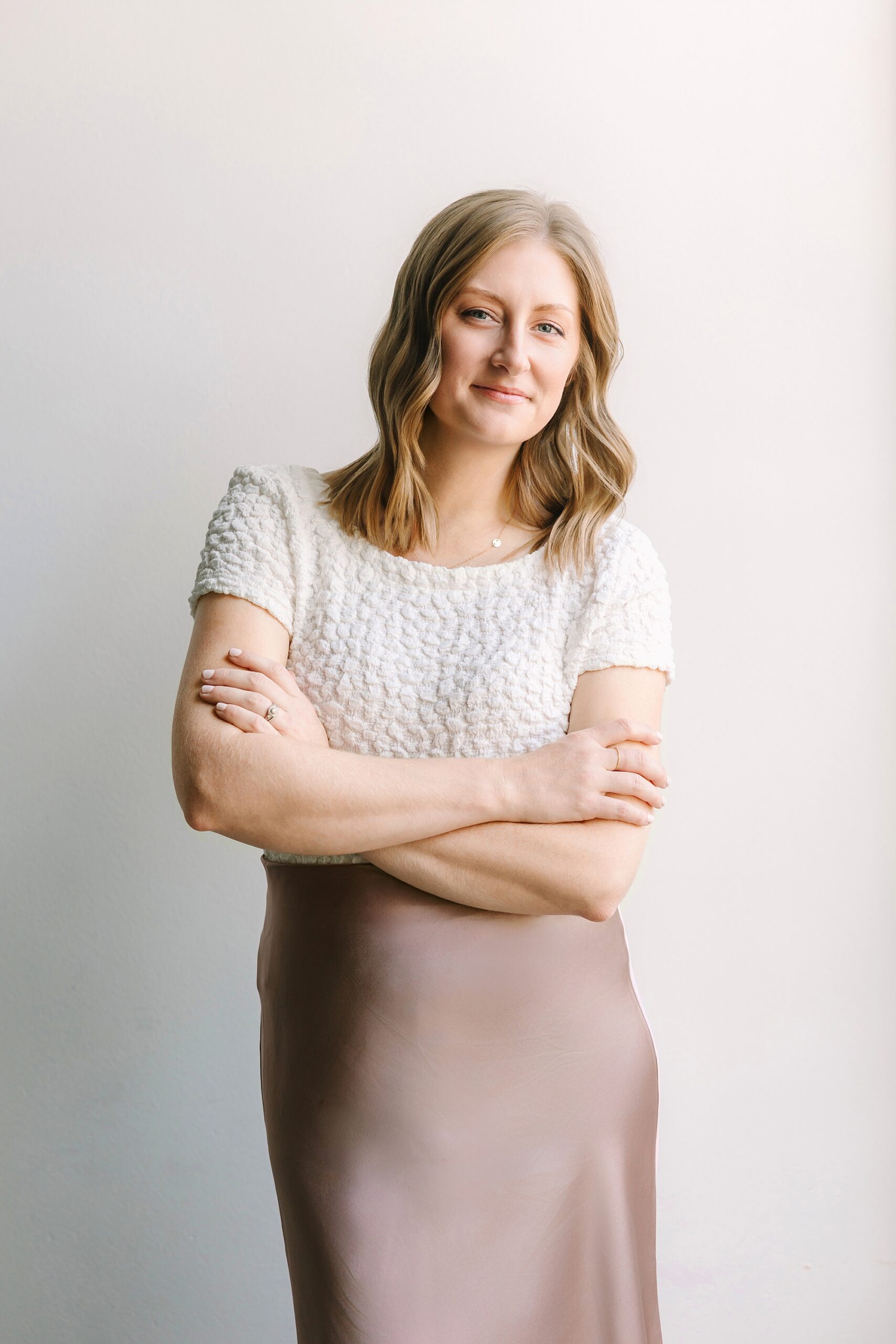 Headshot of Woman with blonde hair in a white top and taupe skirt stands with arms crossed, smiling at the camera.