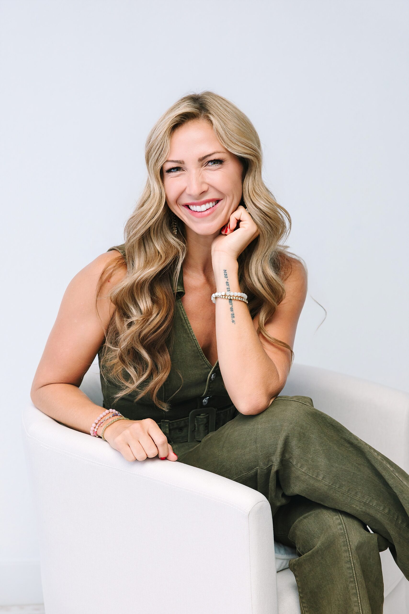 Woman with long blonde hair in a green jumpsuit sits smiling on a white chair against a plain light background.