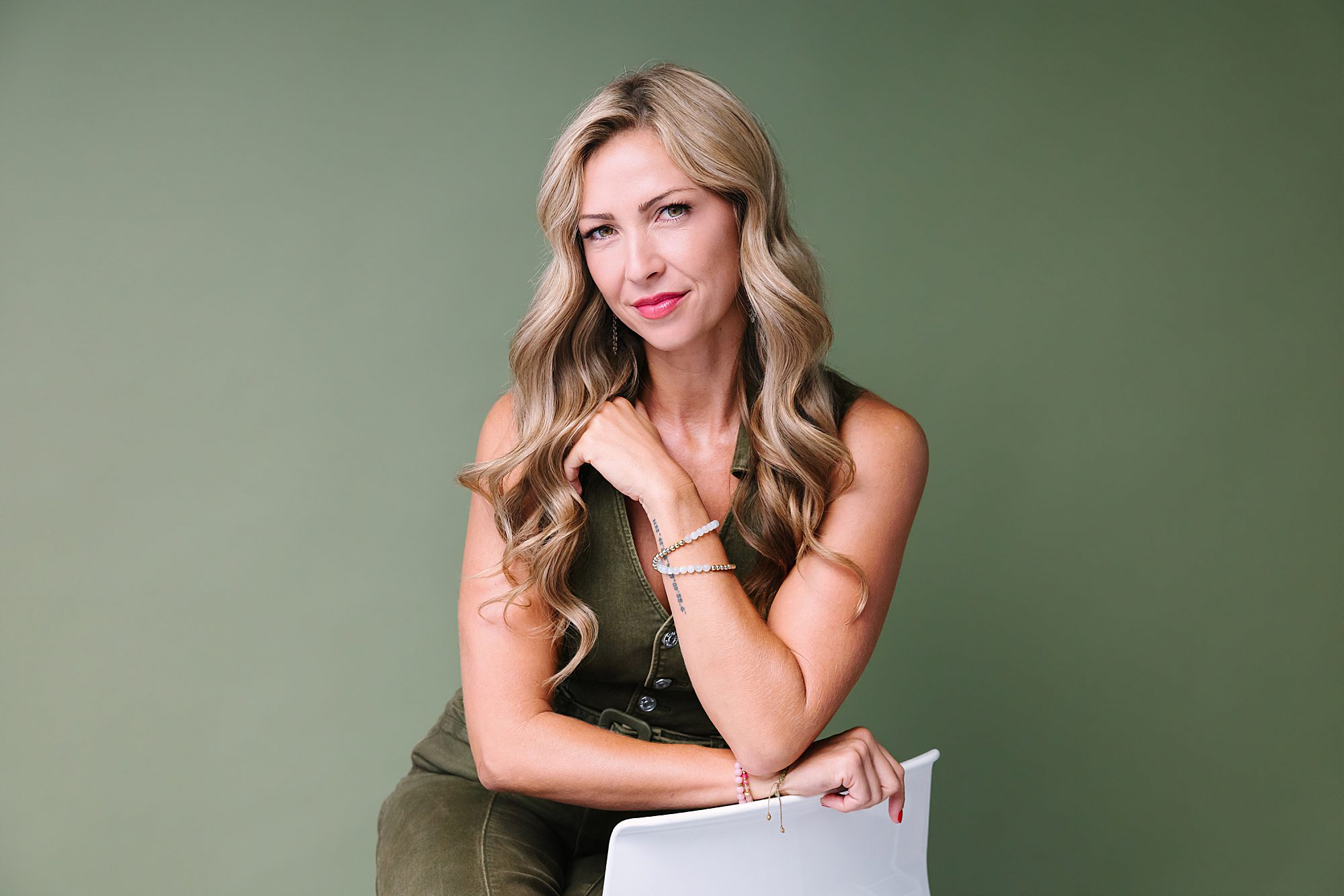 Headshot of Woman with long blonde hair in a green sleeveless outfit sits on a white chair against a green background.