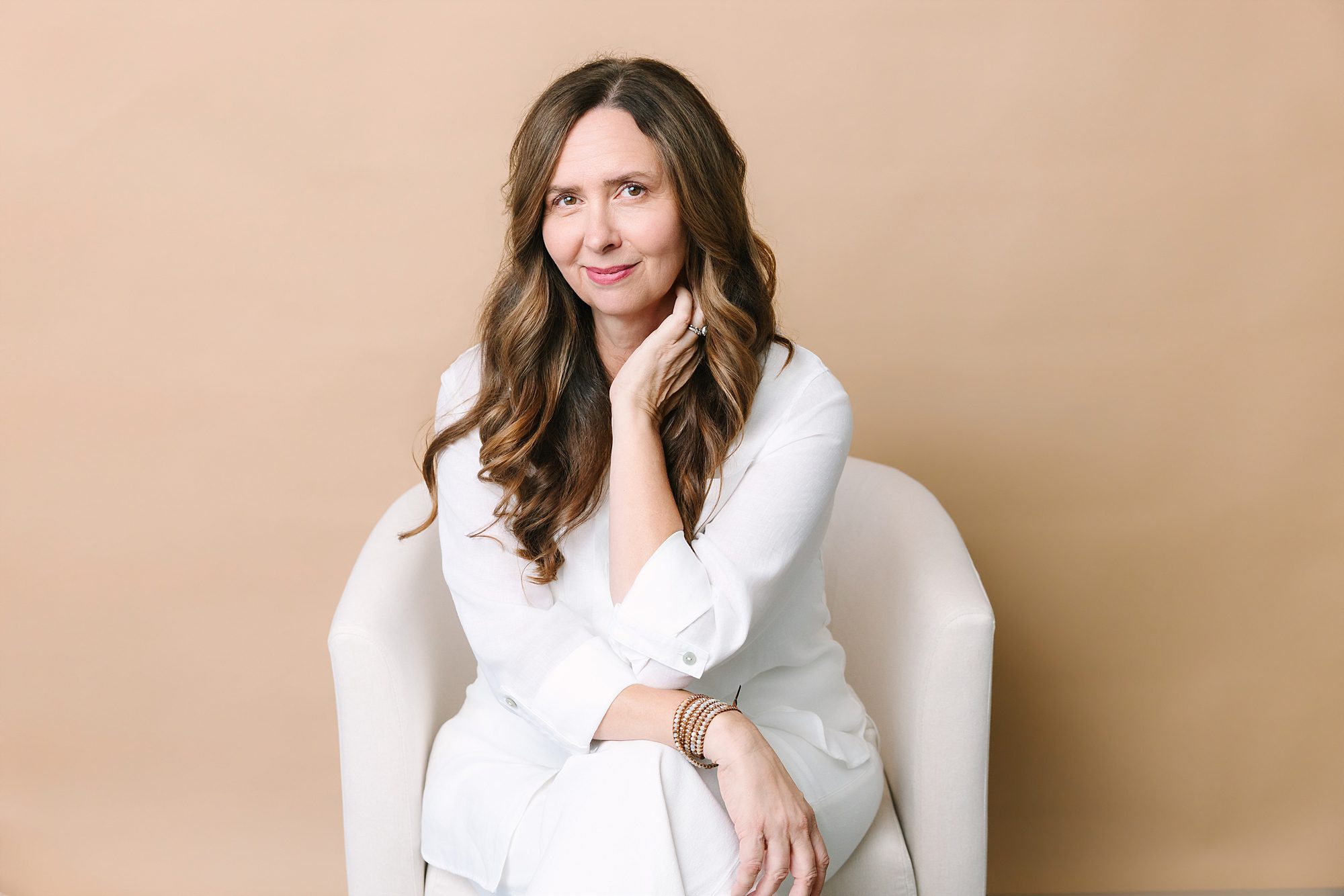 Headshot of Woman with long brown hair in white clothes sits in a chair, smiling, with a beige background.