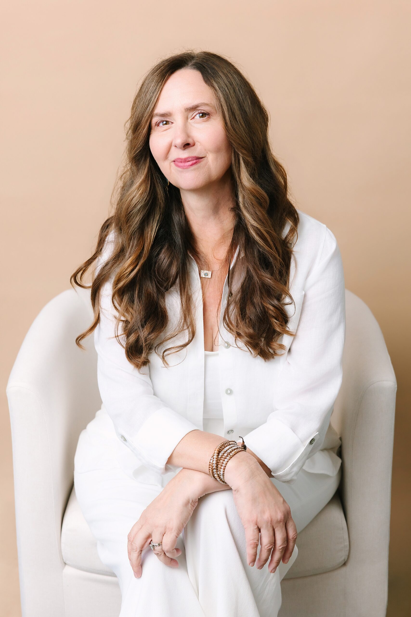 Headshot of Woman with long brown hair in white outfit sits in a cream chair, smiling softly against a beige background.