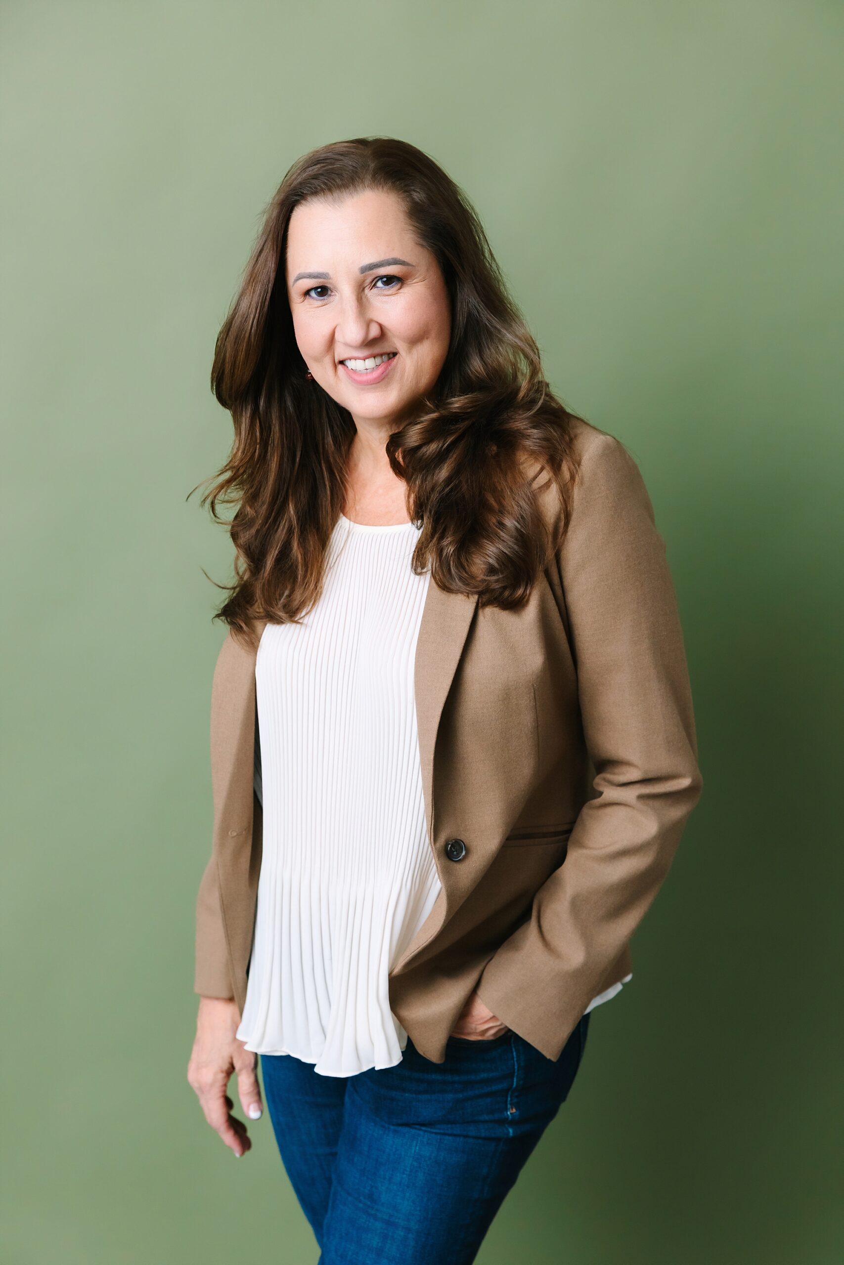 Headshot of Woman with long brown hair, wearing a tan blazer and white blouse, smiles in front of a plain green background.