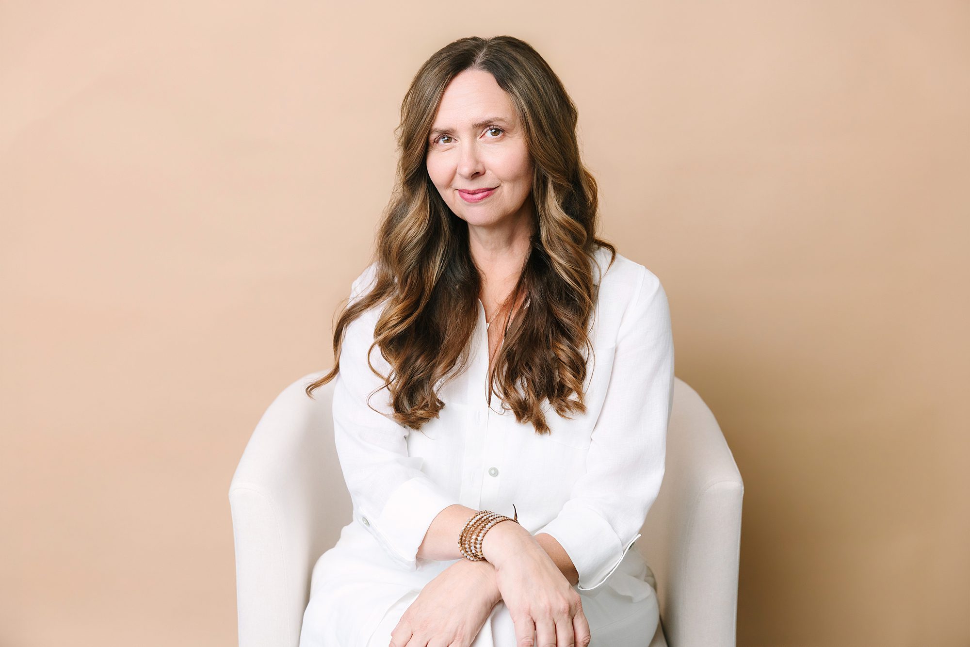 Headshot of Woman with long wavy hair in a white outfit sitting in a white chair, posing against a beige background.