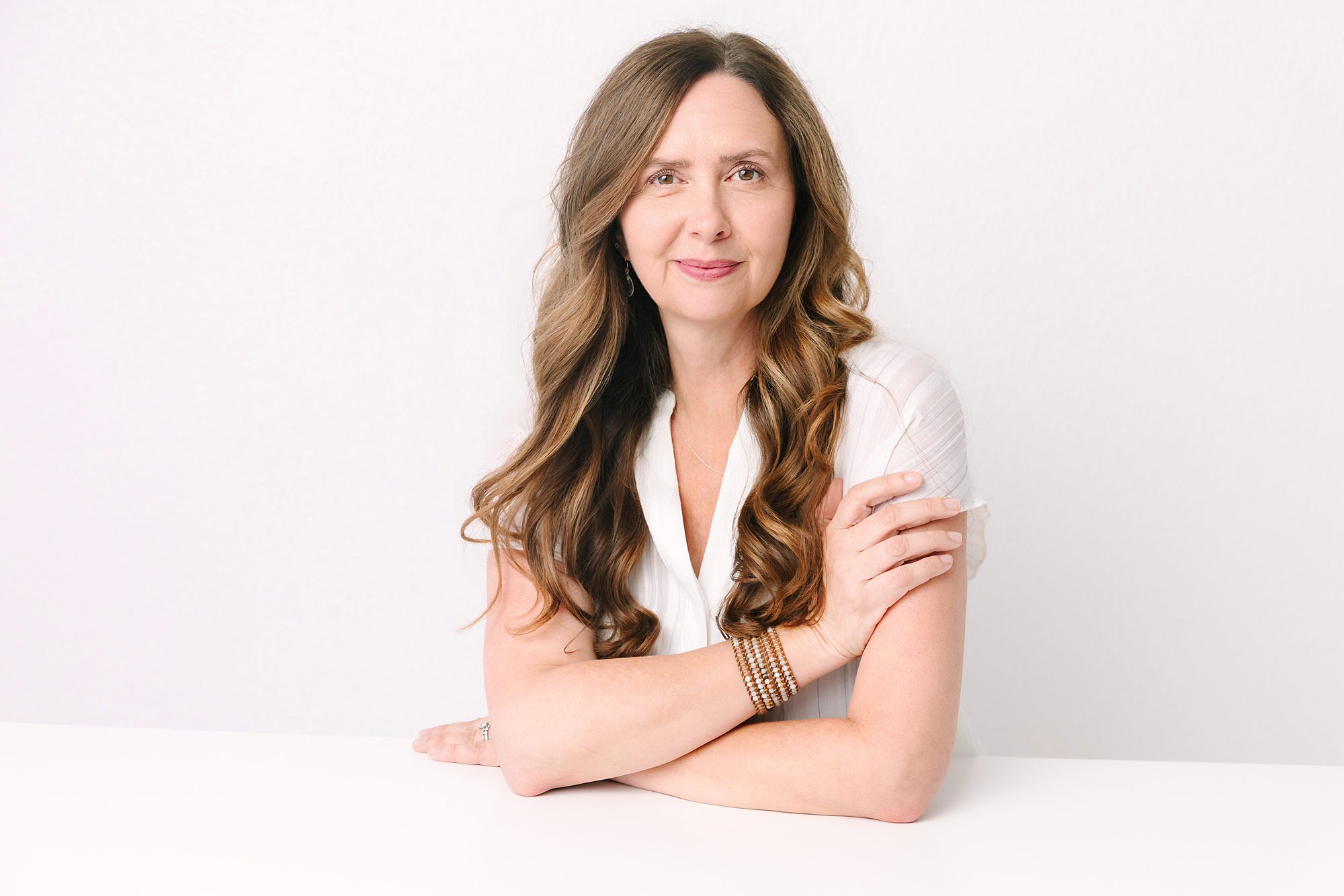 Headshot of Woman with long wavy hair in a white shirt sits at a table, smiling slightly, against a plain white background.
