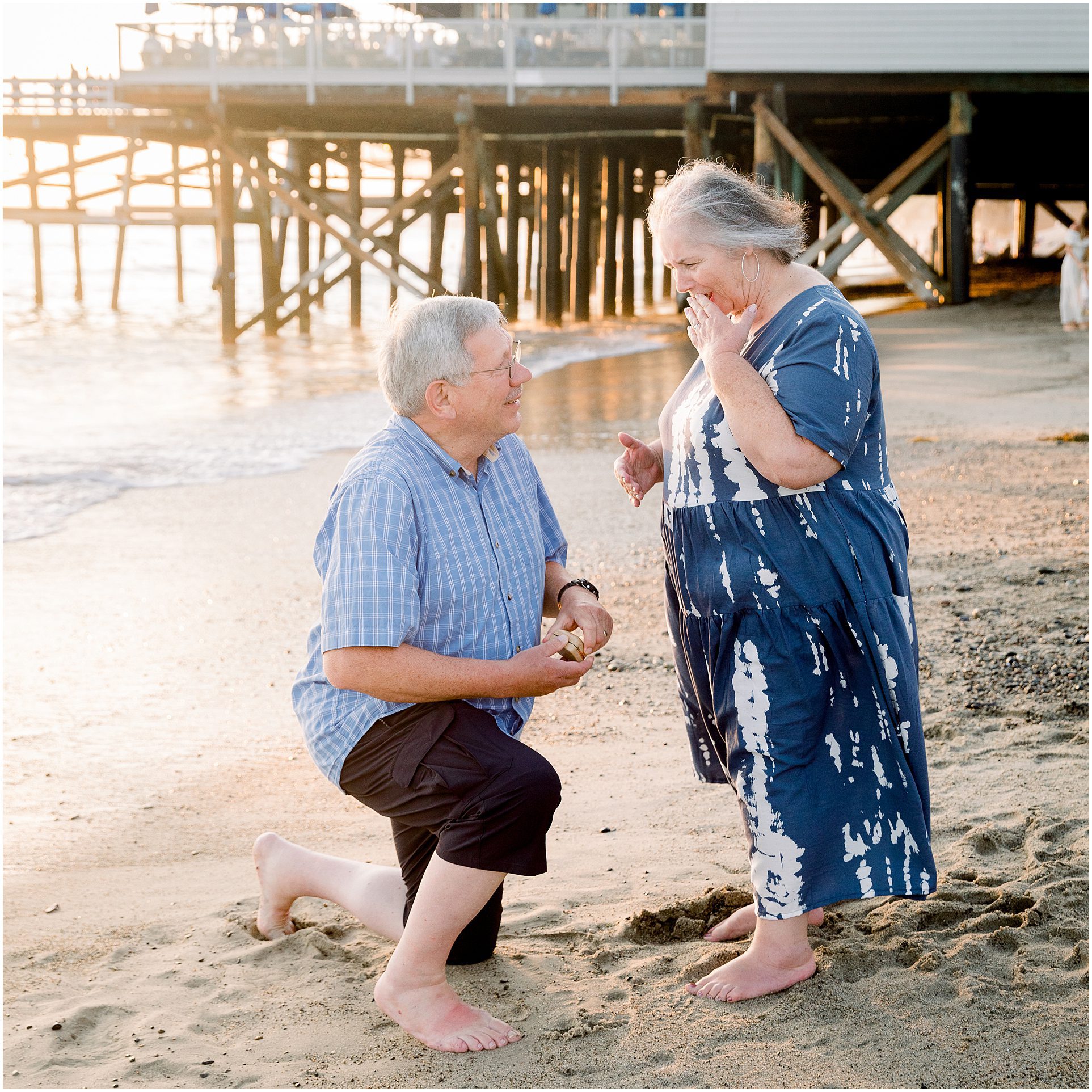 An older man kneels and proposes to an older woman on a sandy beach near a pier at sunset.