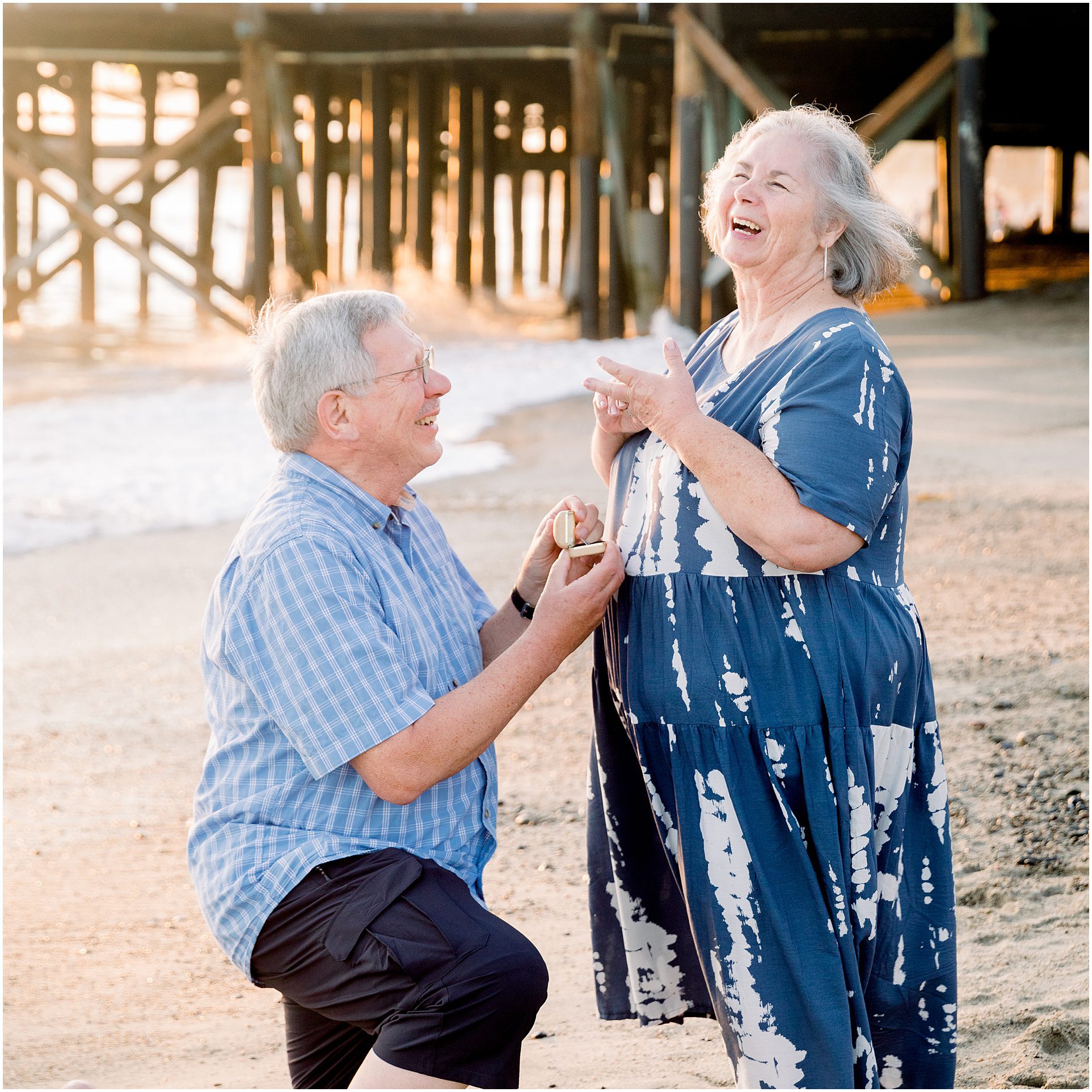 An older man proposes to a joyful woman on a beach near a pier at sunset.