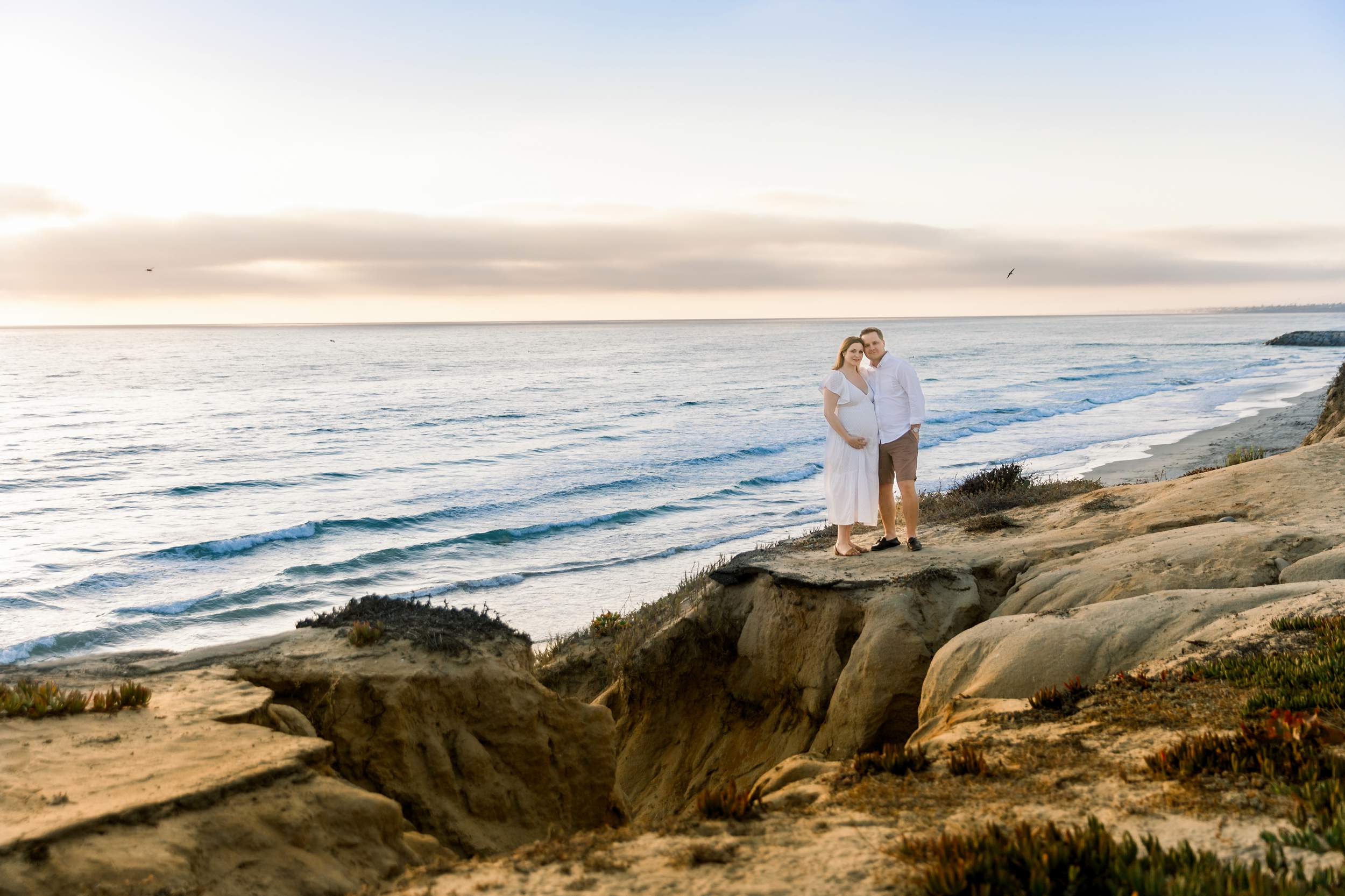 A couple stands on a cliff overlooking the ocean at sunset, with waves and sandy cliffs in the background.