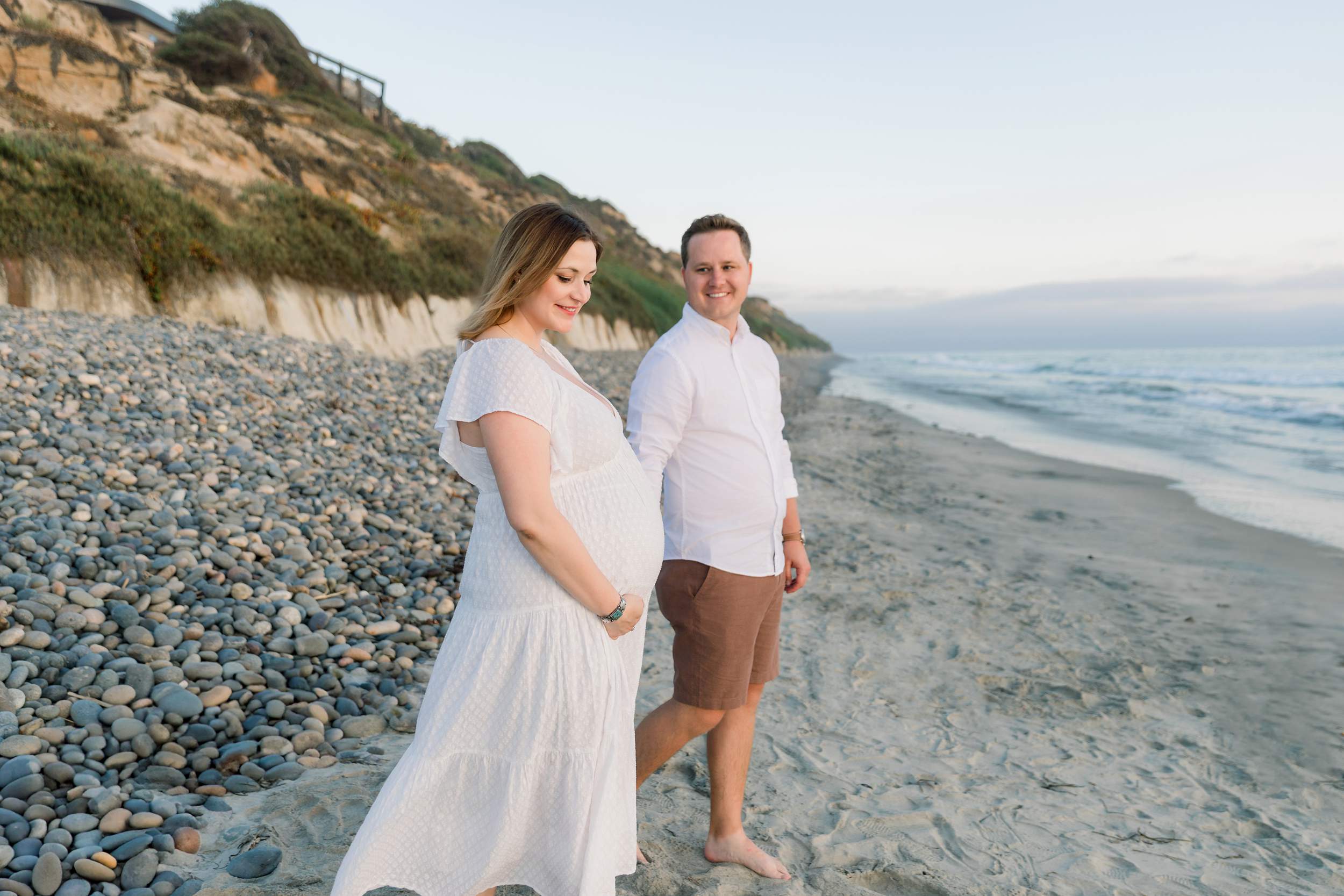 A pregnant woman and a man walk barefoot on a sandy beach near rocks, holding hands and smiling.