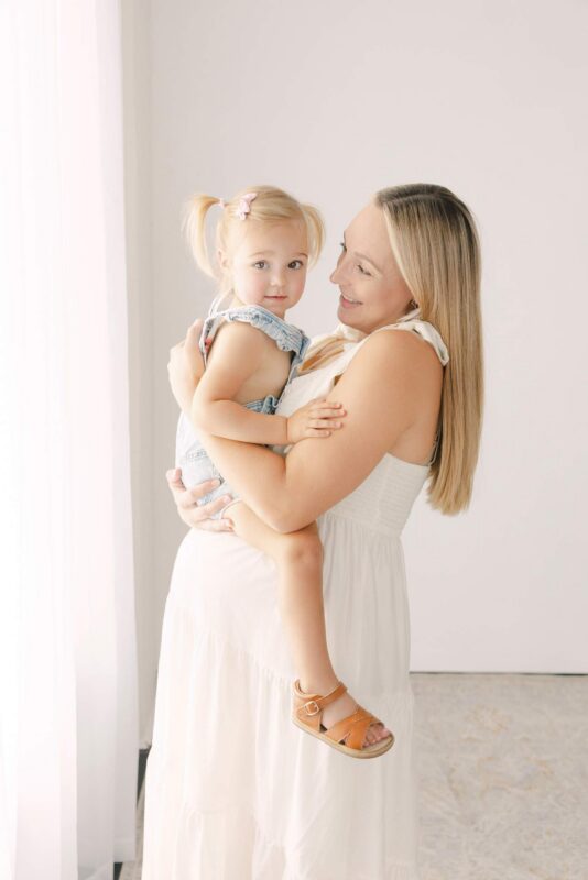 A woman in a white dress holds a young girl with pigtails, both smiling in a softly lit room.