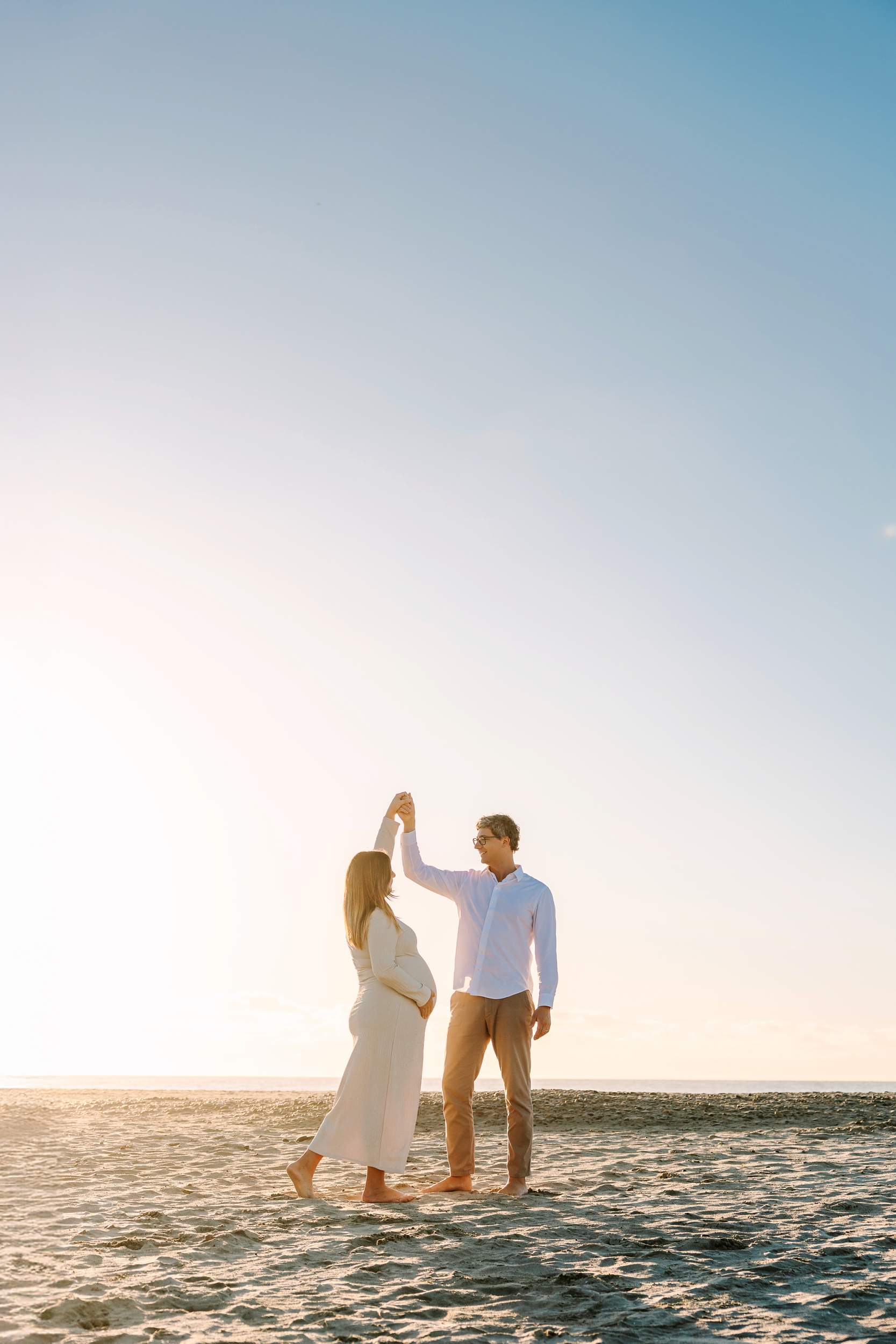 A couple dances on a sandy beach at sunset, with the woman appearing pregnant and the sky clear above them.