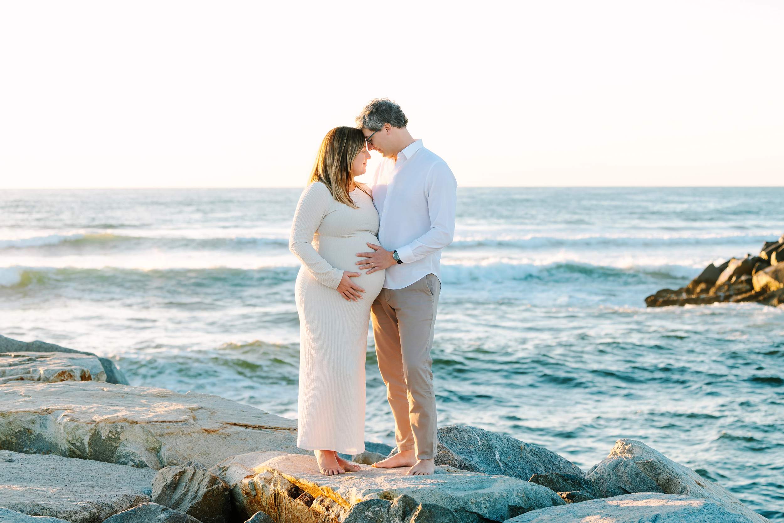 Pregnant woman and man stand barefoot on rocks by the ocean, holding her belly during a maternity photo shoot.