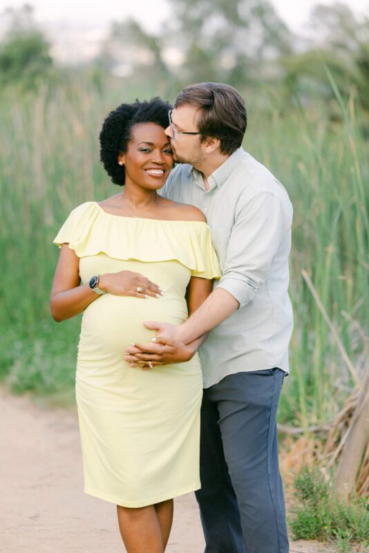 photo by Vallentyne Photography of A pregnant woman in a yellow dress stands outdoors, smiling as her partner hugs her from behind and kisses her on the temple. They both have their hands on her belly, surrounded by tall grass.