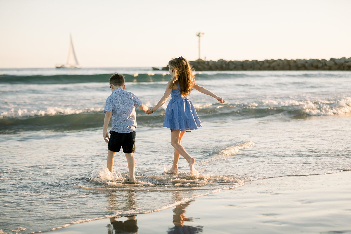 Two children holding hands and walking in shallow water at the beach, with waves and a sailboat in the background.
