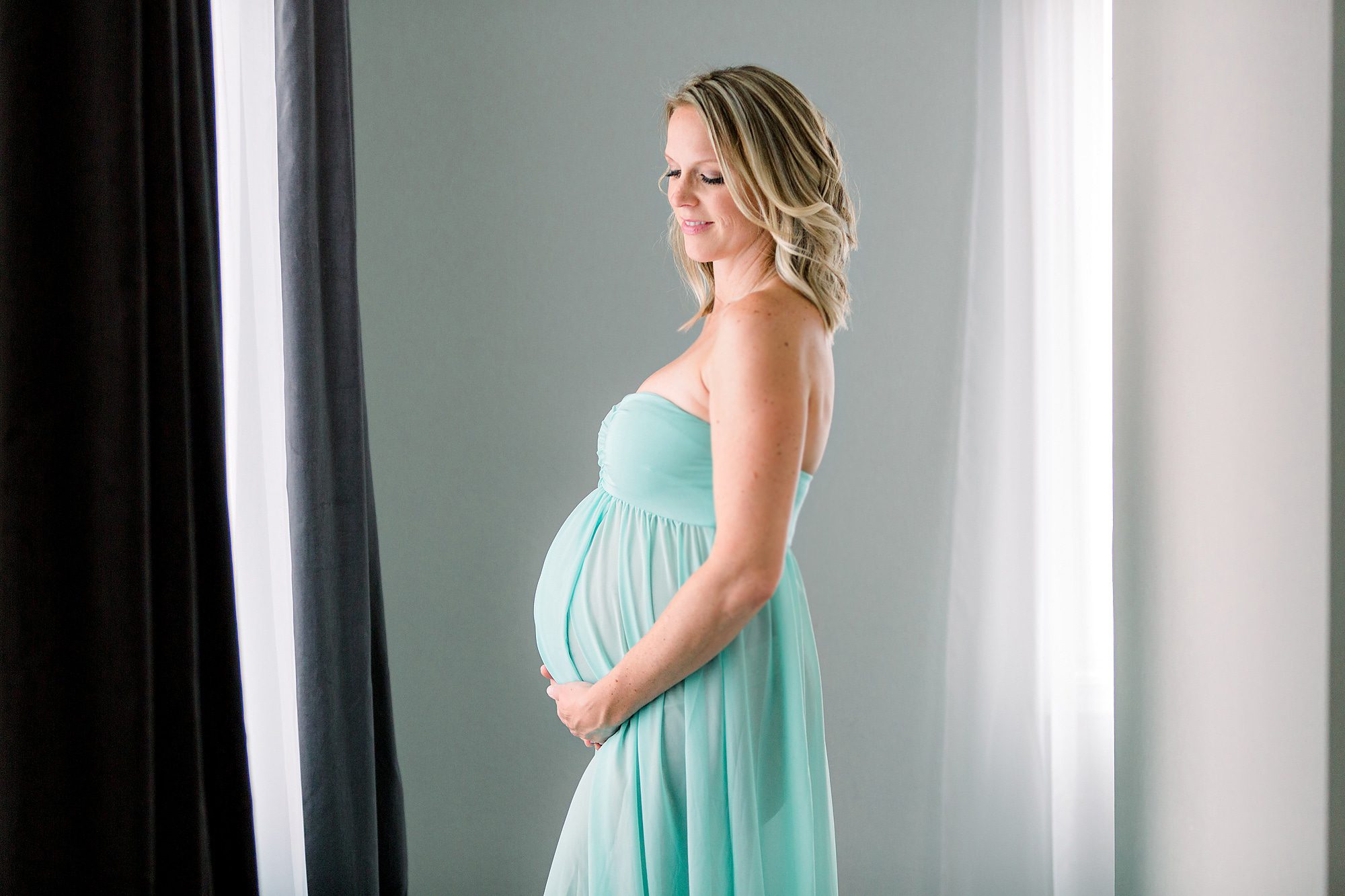 A mother to be in a teal maternity gown stands in a window smiling down at her bump in her hands San Diego Birthing Centers