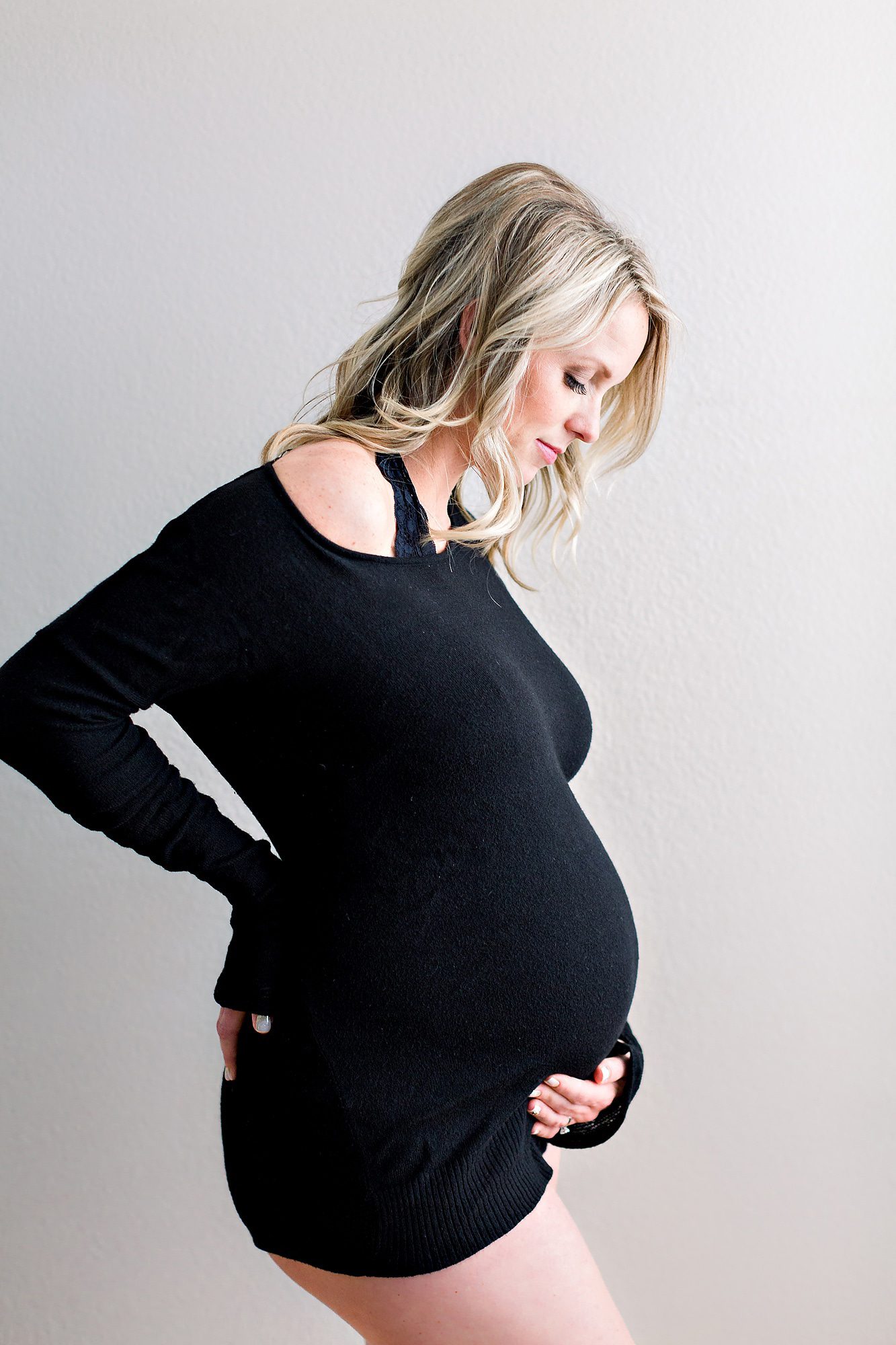 A mom to be in a black long sleeve stands in a studio smiling down at her bump after visiting San Diego Birthing Centers