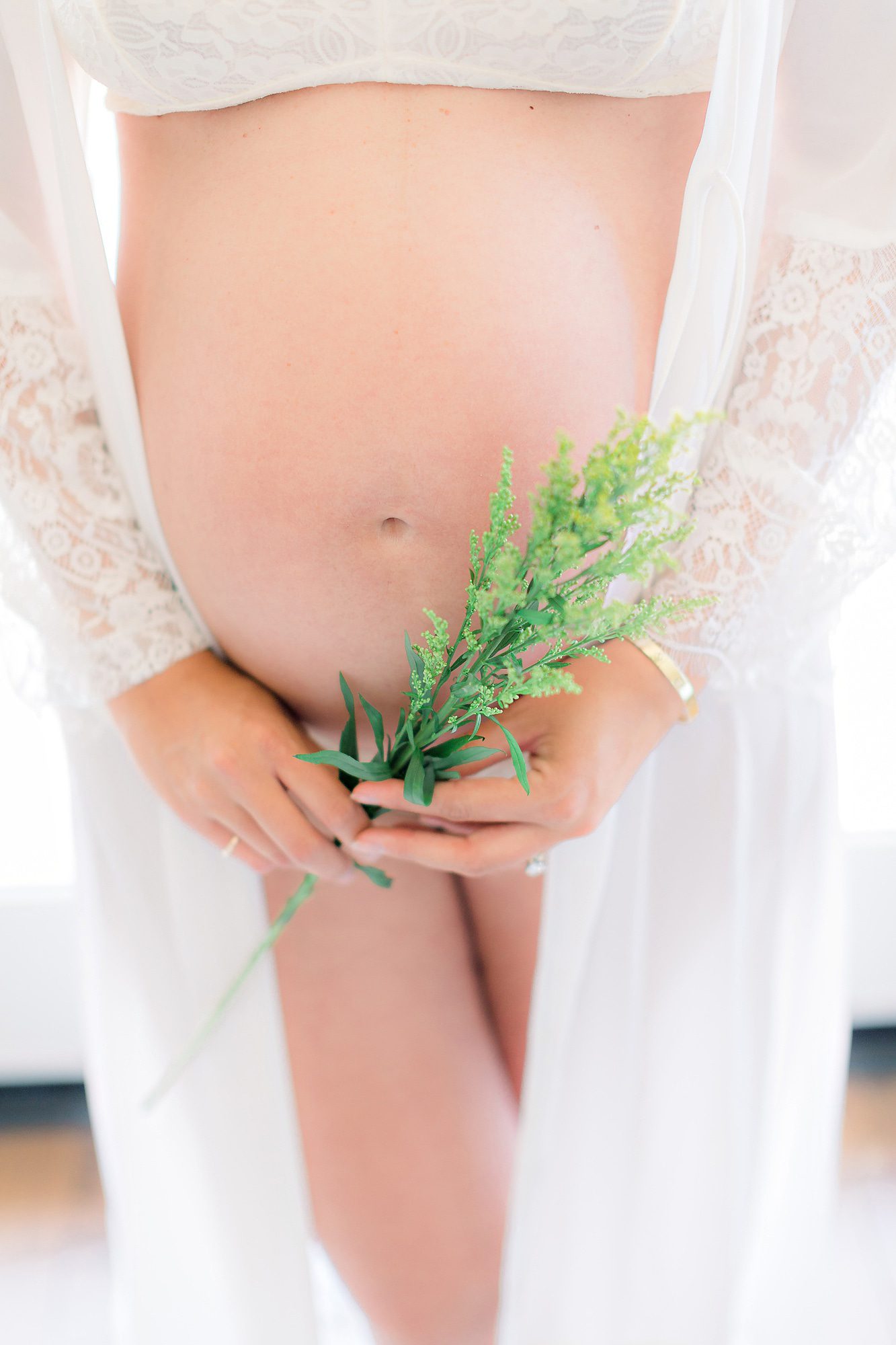 A mother to be standing in a lace coverup with bump out and holding some greenery after visiting San Diego Birthing Centers