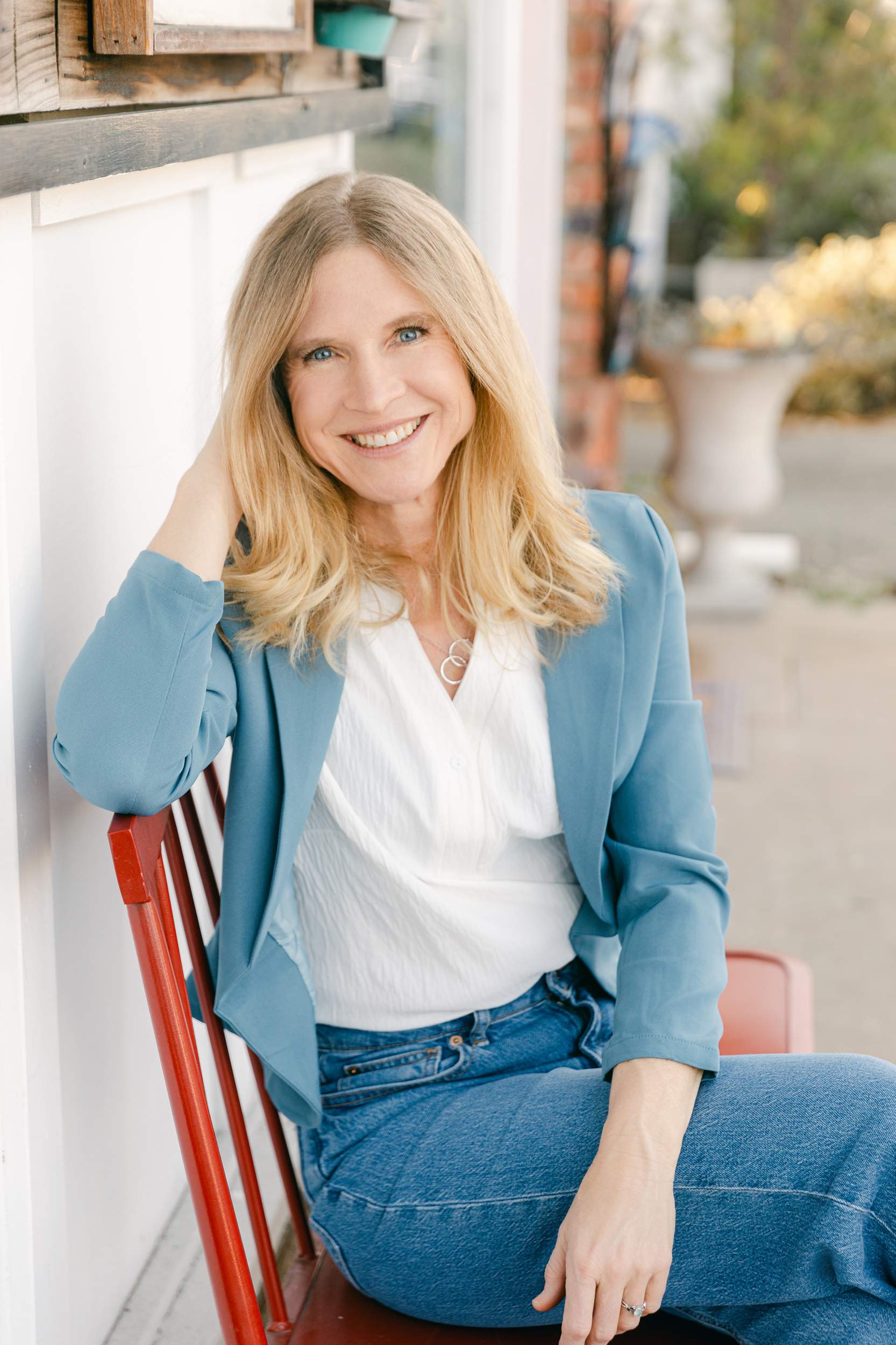 photo by Vallentyne Photography of A smiling woman with long blonde hair sits on a red chair outdoors, styled in a light blue blazer, white blouse, and jeans—captured by a personal branding photographer San Diego for an authentic lifestyle portrait.